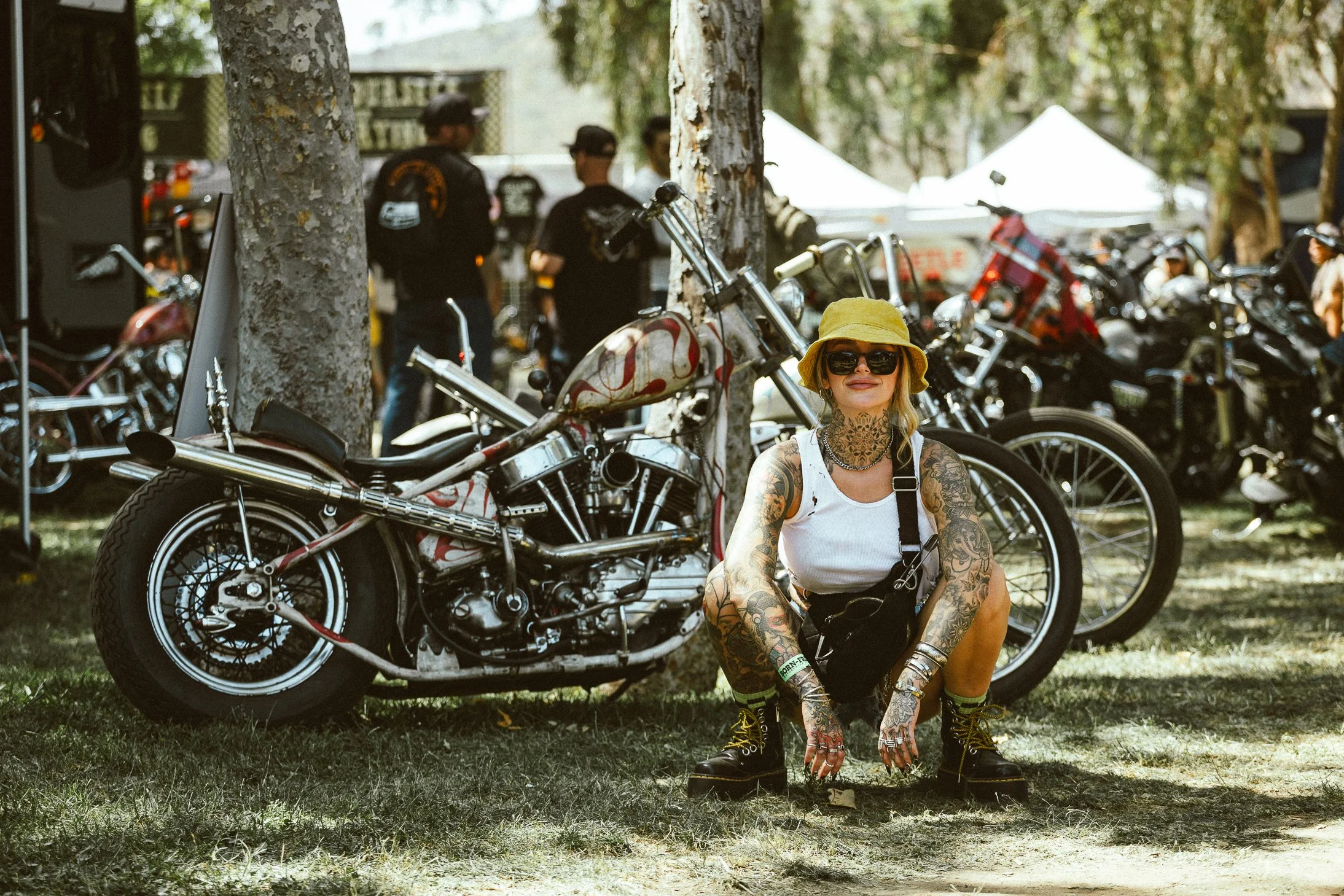 A woman with blonde hair, tattoos, and wearing sunglasses, a yellow hat, a white tank top, and combat boots, crouches next to a vintage motorcycle at an outdoor event with many motorcycles and tents in the background.