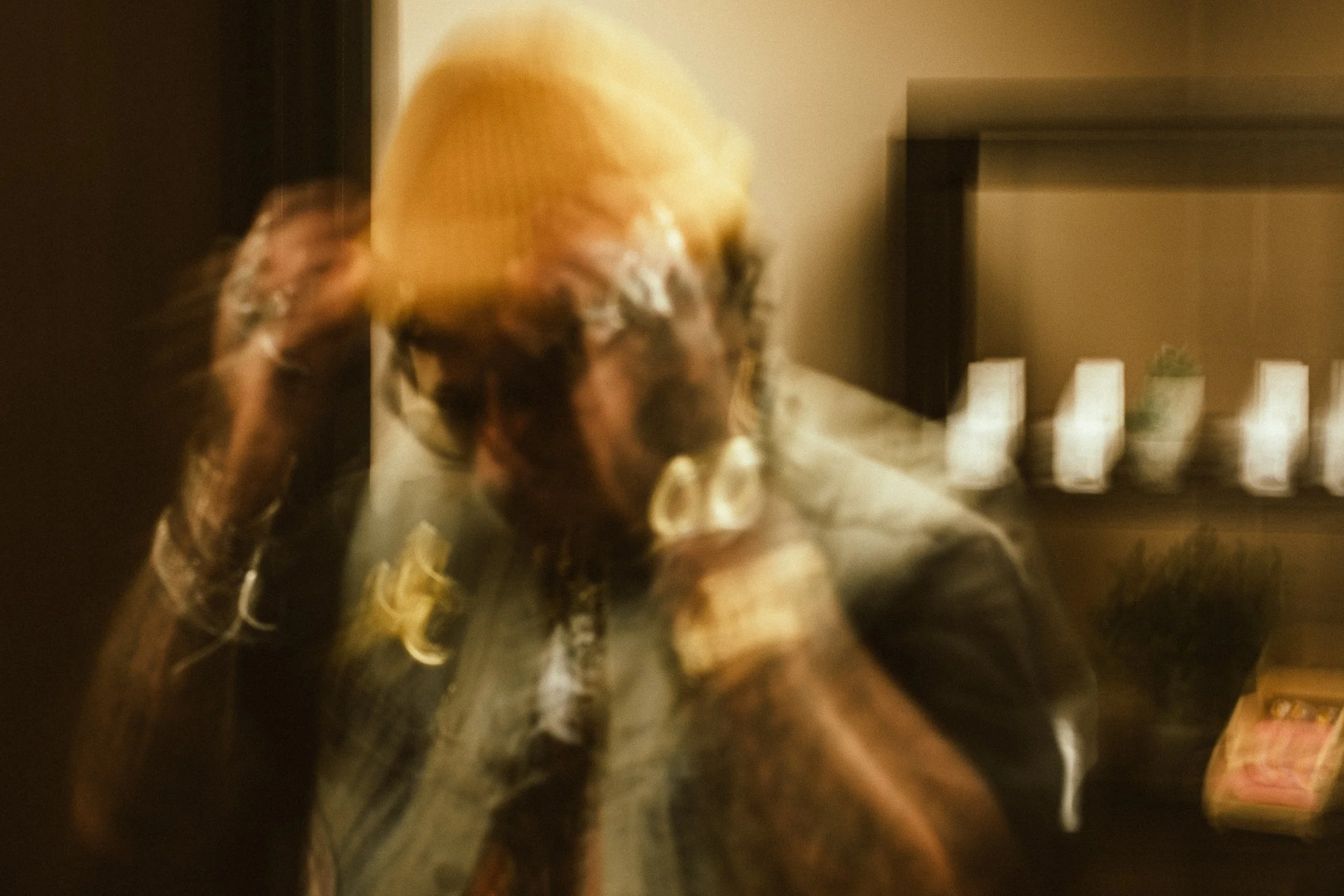 Blurry photo of a person with long hair and sunglasses, wearing an orange beanie, sitting at a desk with a computer monitor and a small pink box, in an indoor setting with warm lighting.