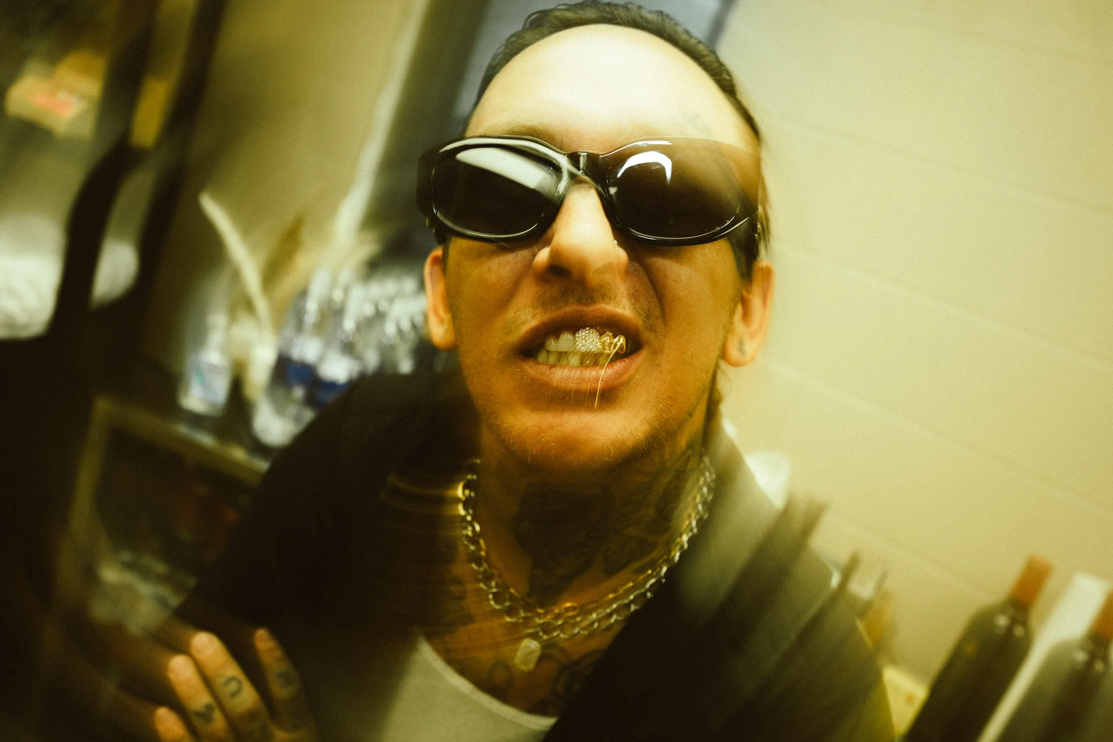 Man with sunglasses showing gold teeth and multiple necklaces, leaning forward in a dimly lit room with bottled water in the background.