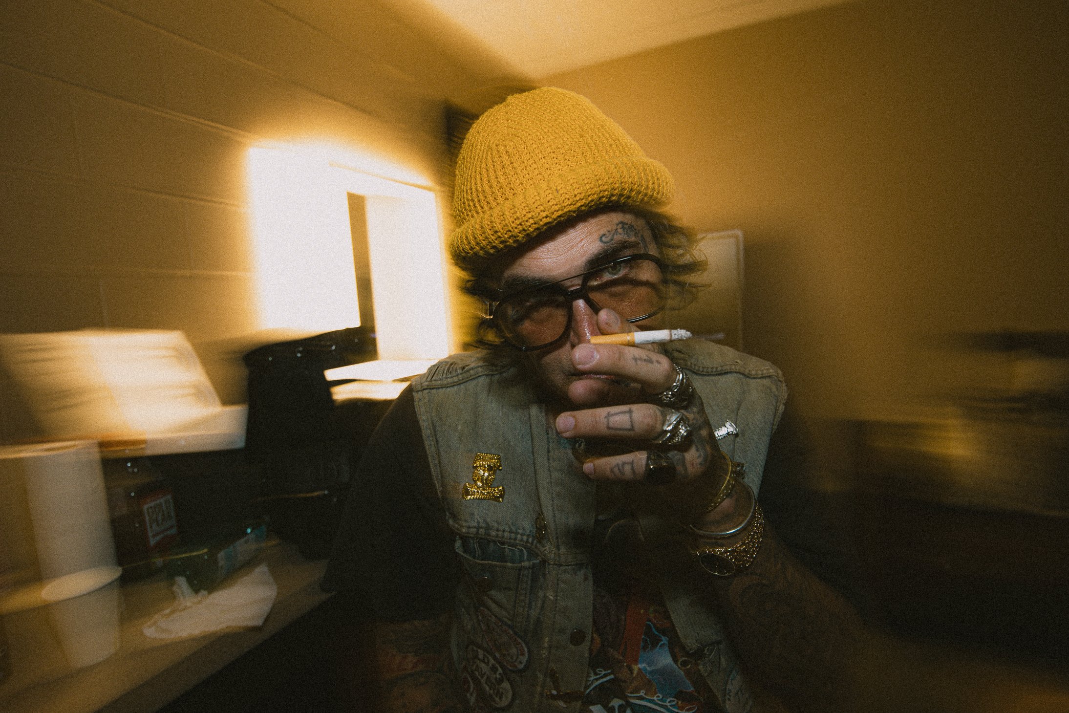 Person wearing a yellow knit hat, sunglasses, and a denim vest, holding a cigarette near their nose and looking at the camera in a dimly lit room with a yellowish background.