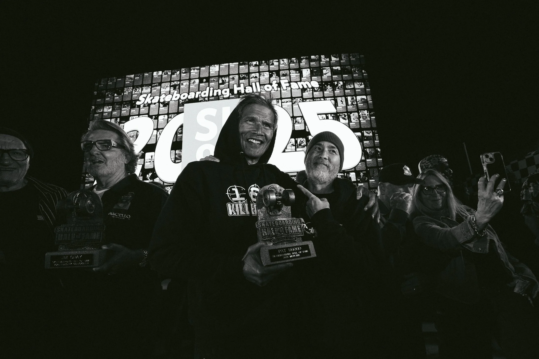 Black and white photo of people celebrating at a skateboarding Hall of Fame event, with two individuals in front holding awards and smiling, behind a large illuminated sign reading "Skateboarding Hall of Fame 2023."