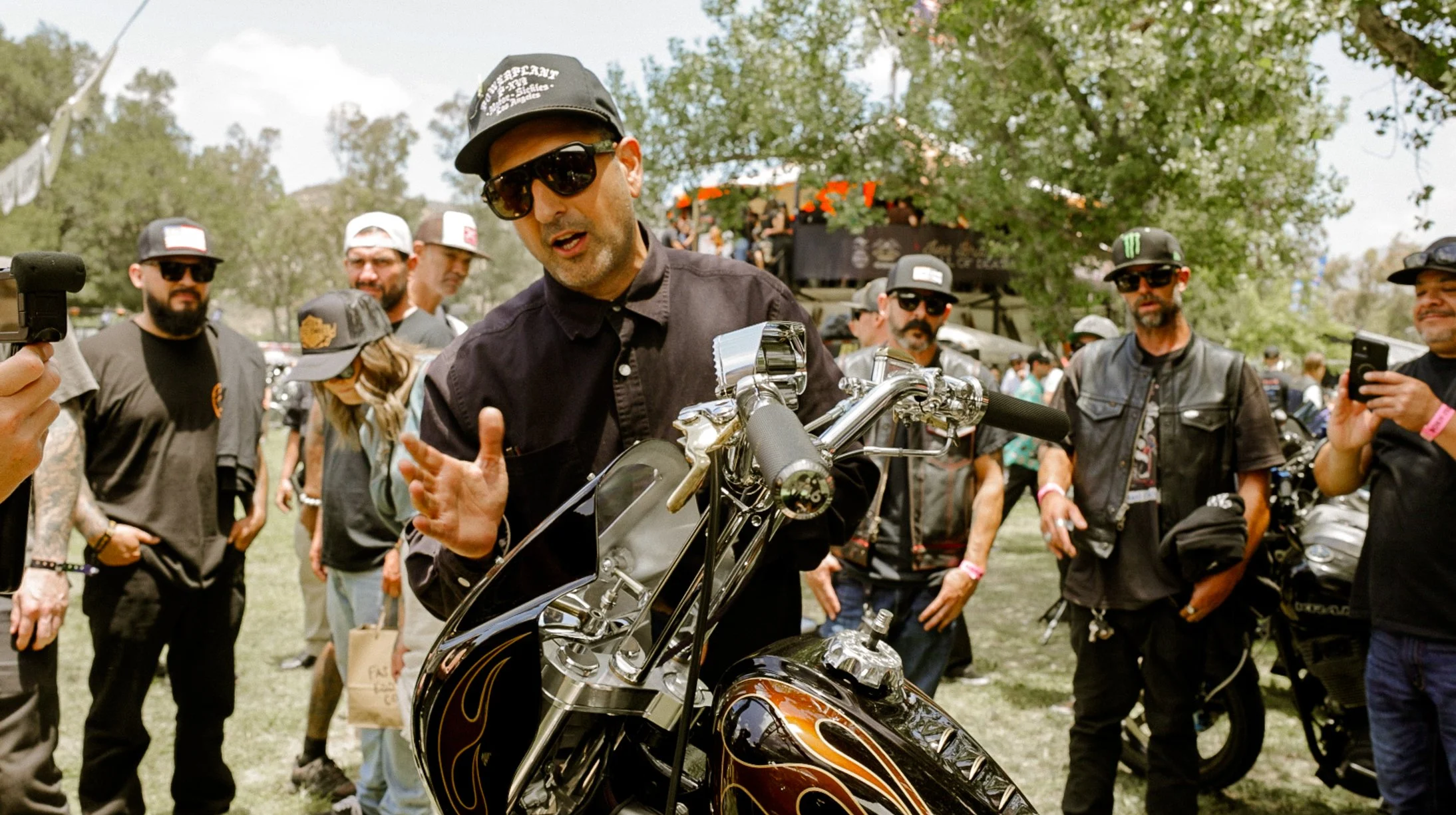 A man in a black shirt and sunglasses is showing a custom motorcycle to a group of people at an outdoor event.
