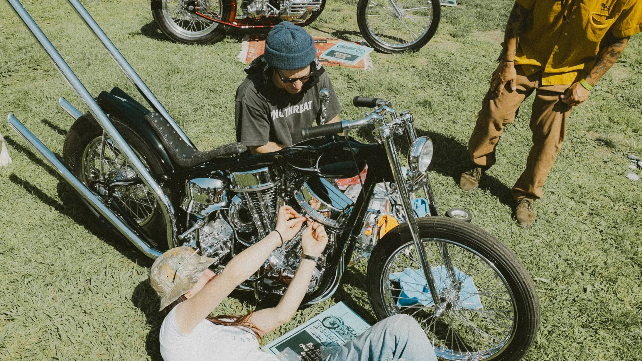Three people working on customizing a black motorcycle with chrome accents on a grassy field, with bicycles in the background.