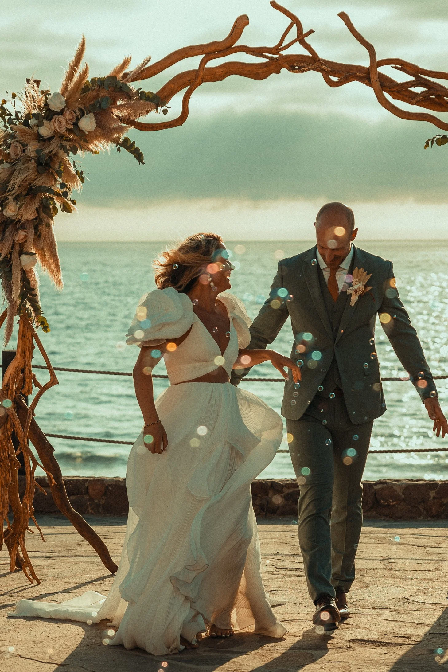 A couple dressed in wedding attire holding hands and dancing on a seaside venue with a decorated arch and ocean background during sunset.