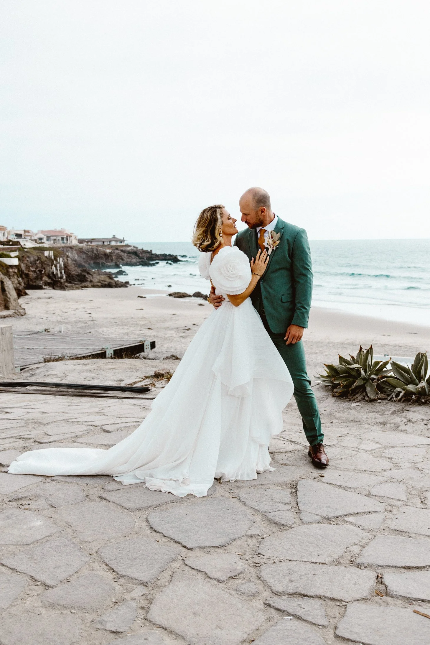 A bride and groom embrace on a beach, with ocean waves and homes in the background during a wedding photoshoot.