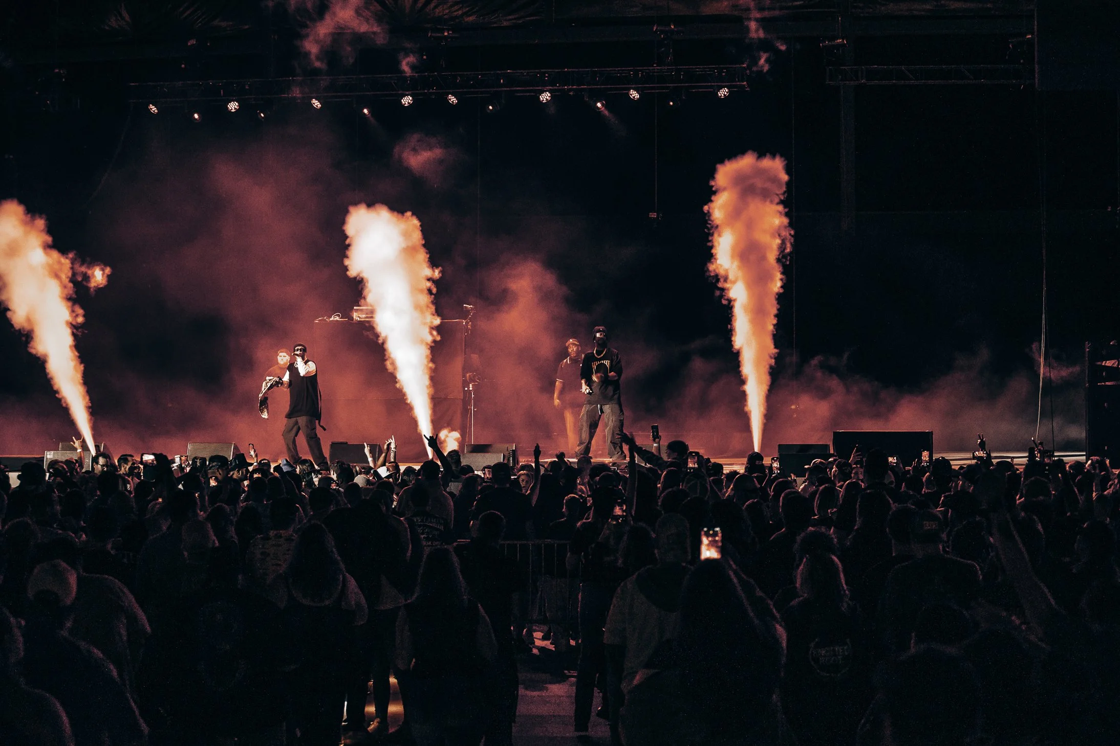 Live concert with multiple performers on stage, pyrotechnics emitting smoke, and a large audience watching.