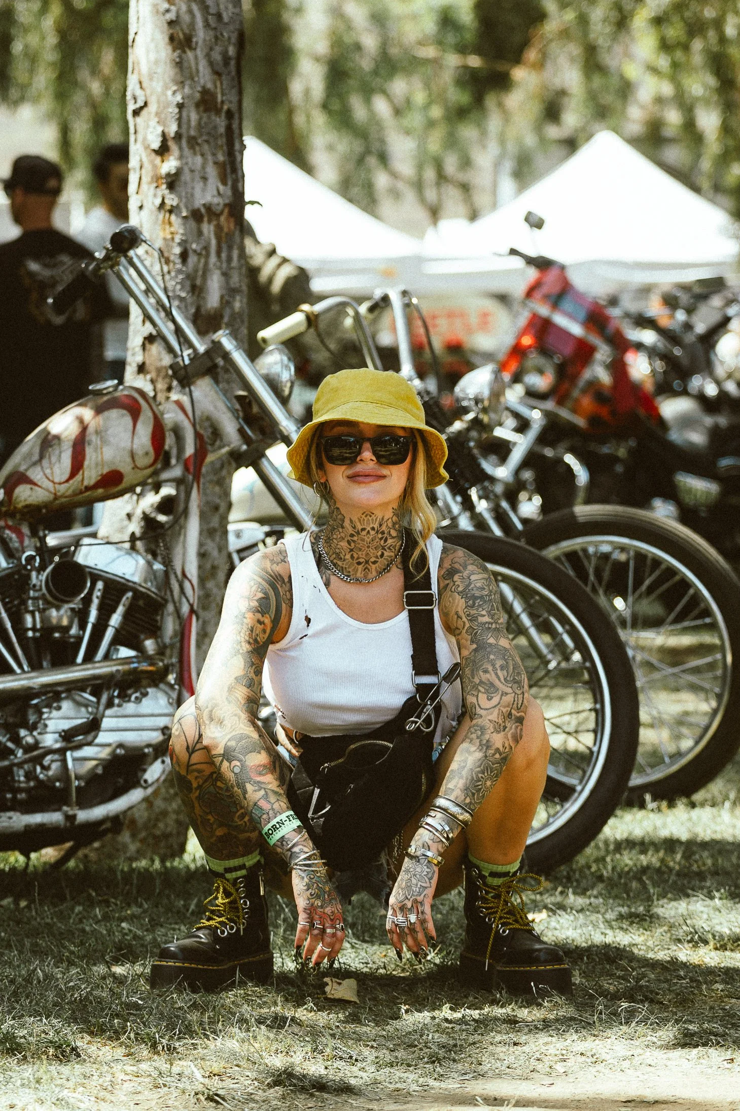 A woman with tattoos wearing a yellow hat, sunglasses, a white tank top, and black boots kneeling on grass at an outdoor motorcycle event with motorcycles and tents in the background.