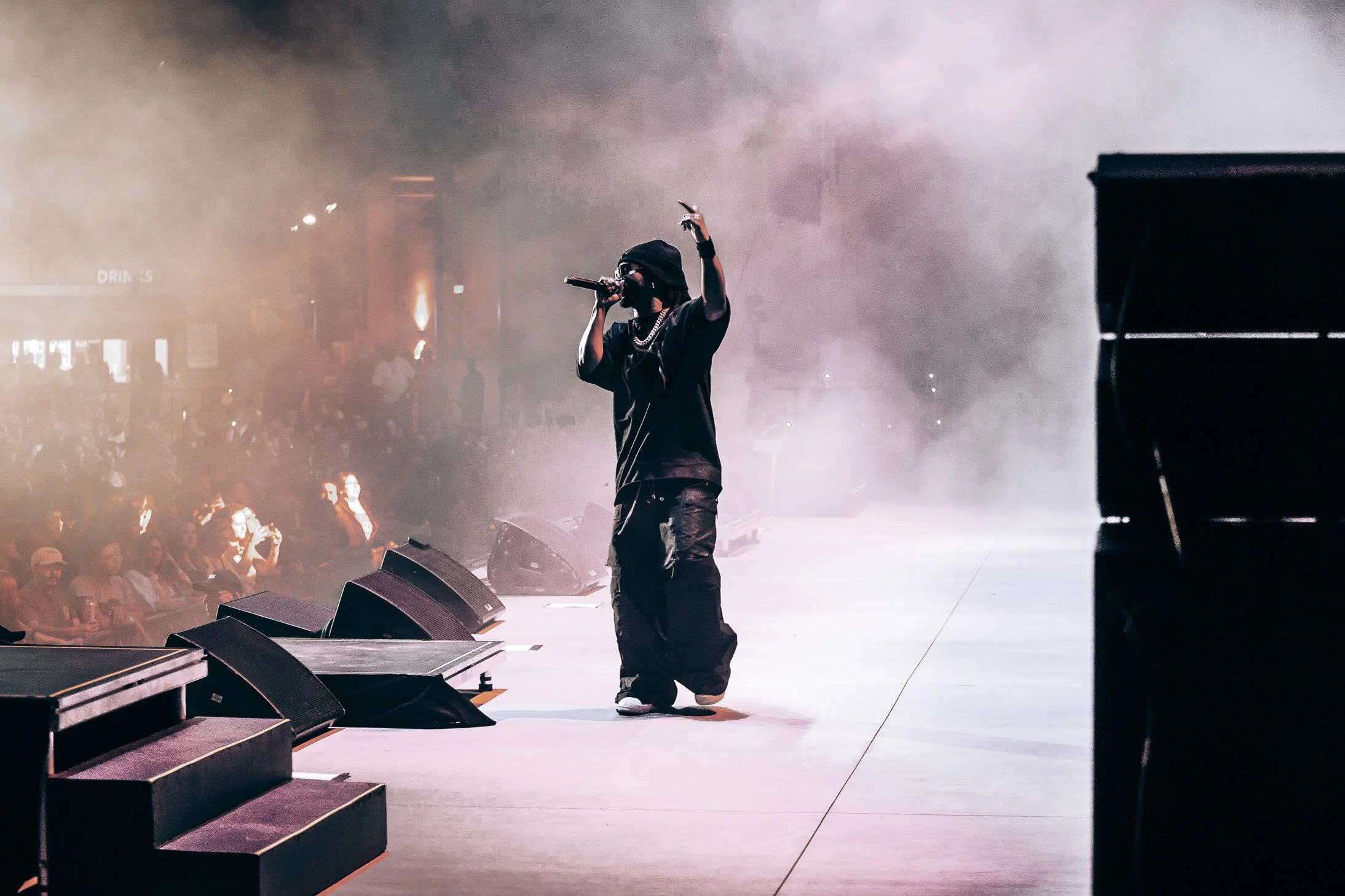 Performer in dark clothing and a black beanie holding a microphone on stage with a smoky background and audience watching.