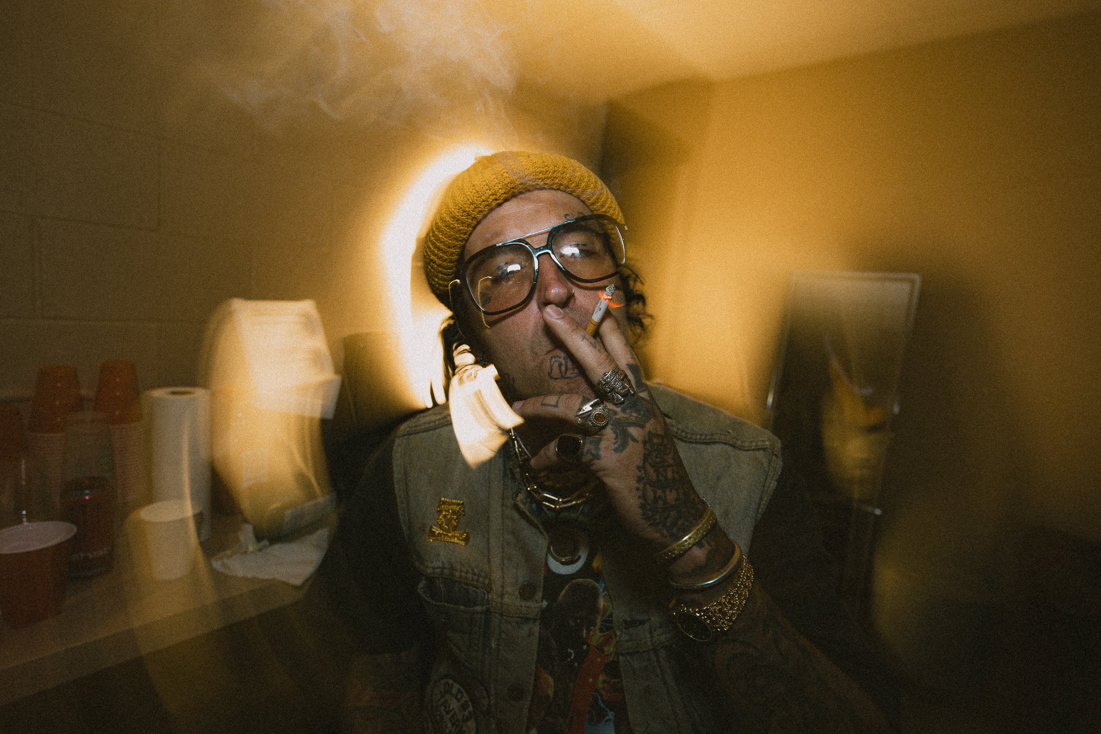 Person with tattoos, wearing glasses and a yellow beanie, smoking a cigarette, in a dimly lit room with a yellow wall, drinks and cups on a table in the background.