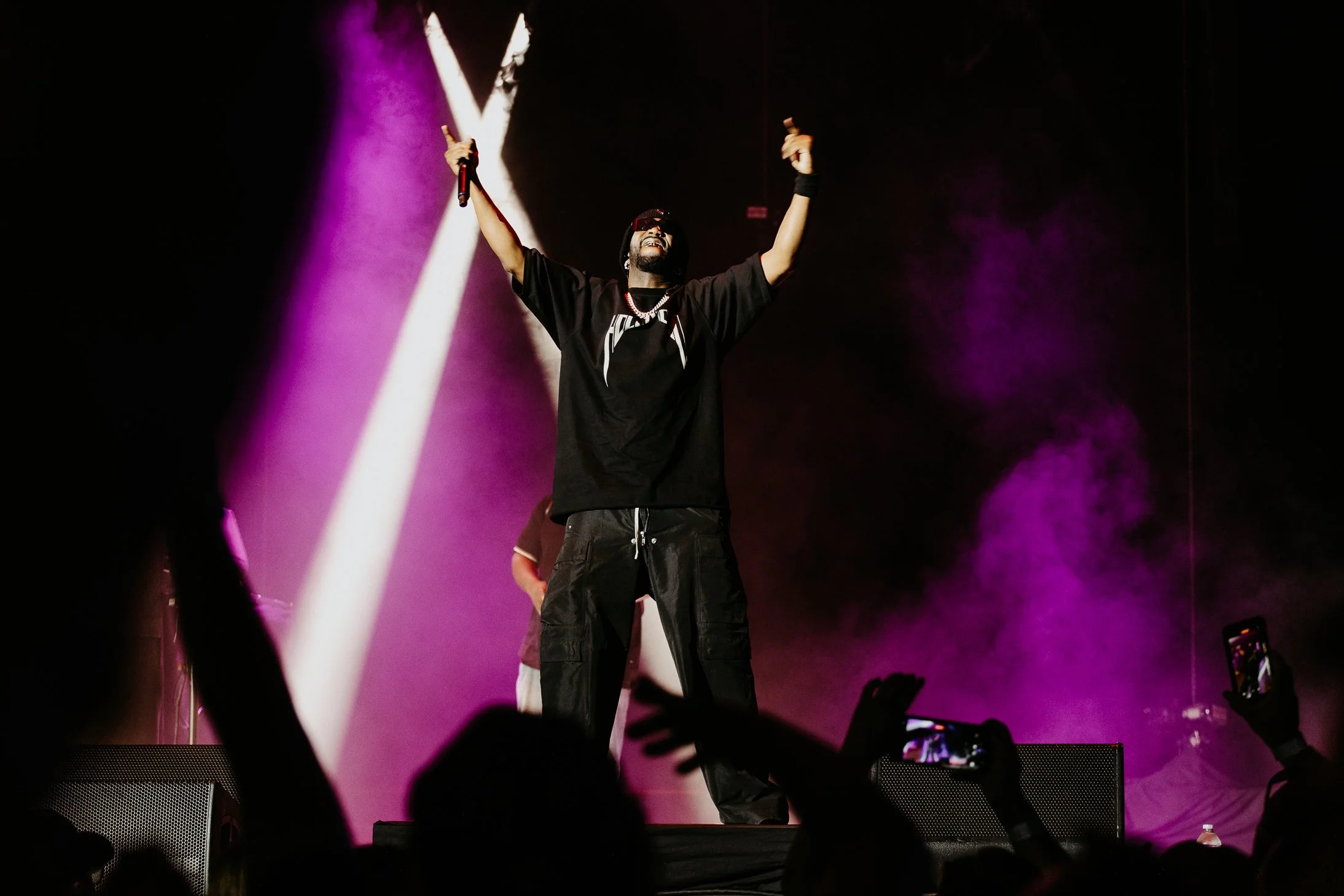 Male performer on stage with arms raised, wearing sunglasses, black shirt, and black pants, illuminated by stage purple and white lights, with audience hands visible in the foreground.