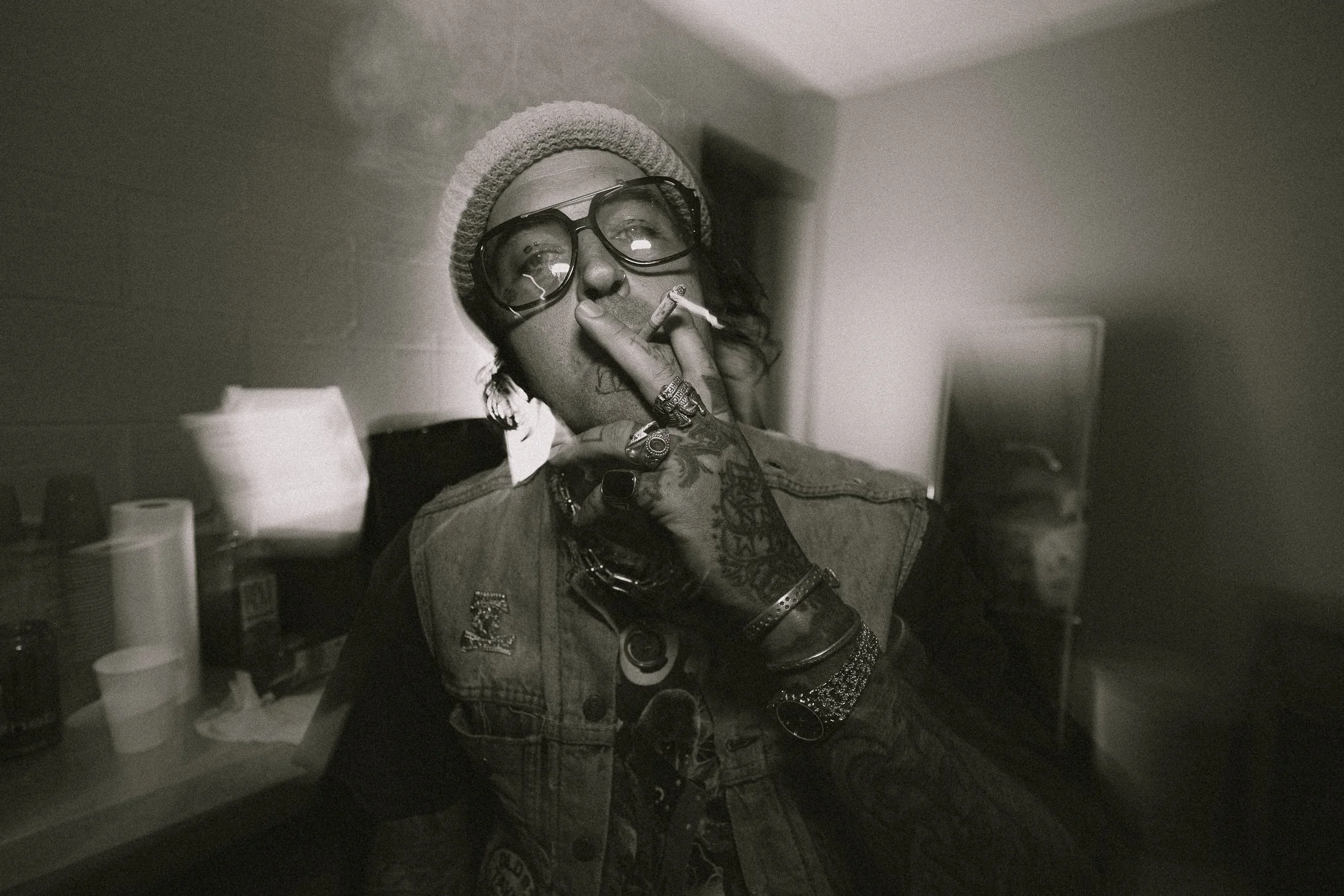 Black and white photo of a person wearing glasses and a beanie, smoking a cigarette, with tattoos on their hand and wrist, in a room with a counter and various objects in the background.