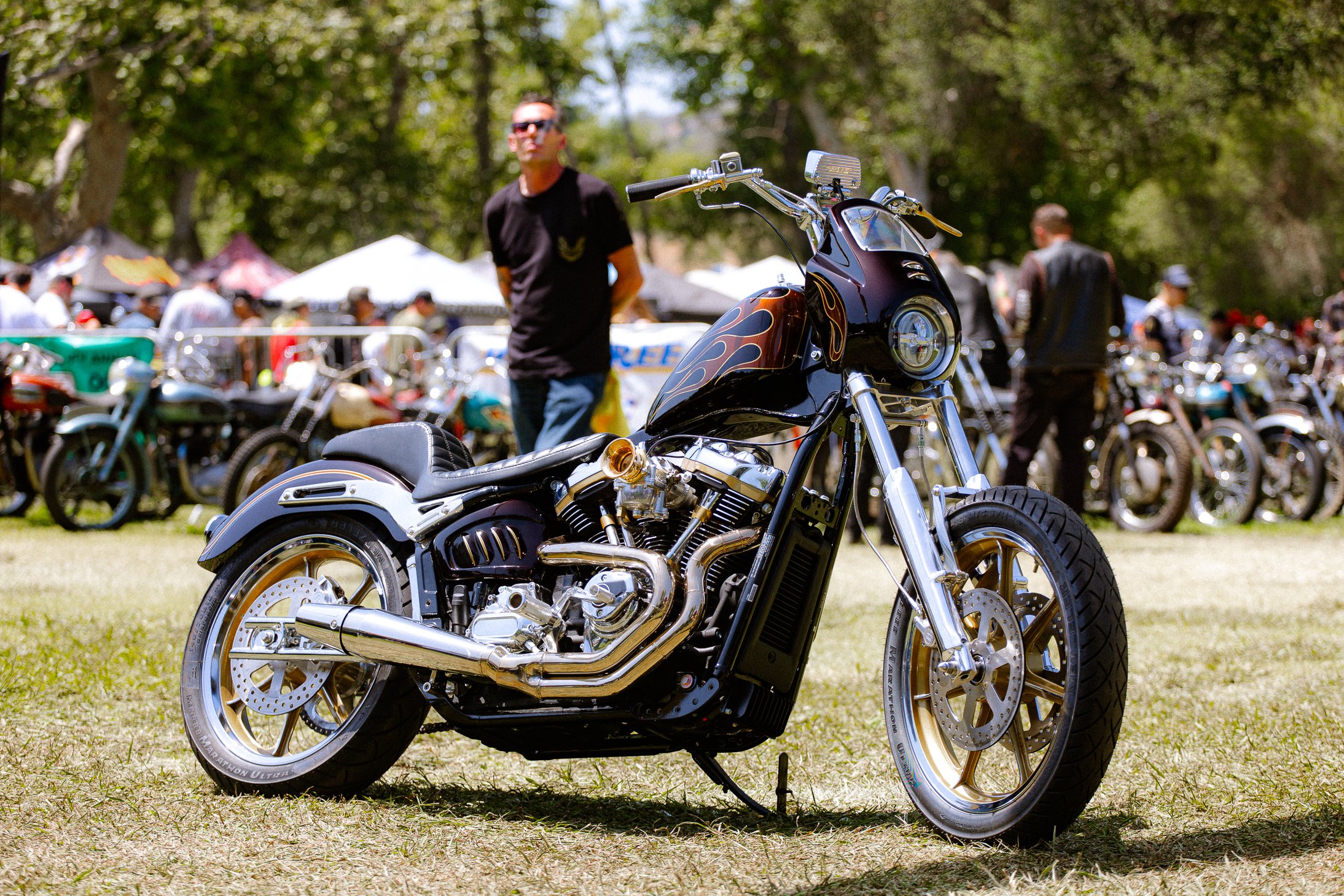 A custom black motorcycle with flames on the gas tank parked on grass at an outdoor show with people and additional motorcycles in the background.