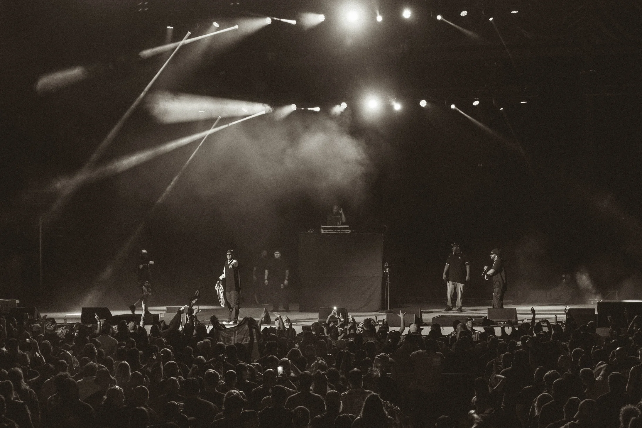 Black and white photo of a large crowd at a concert, with a stage featuring multiple performers and a DJ, colorful lights and smoke effects overhead.