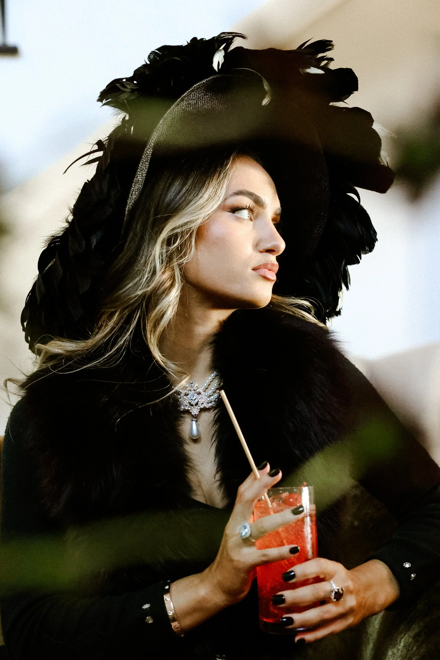 A woman wearing a black feathered hat and a fur coat holds a red cocktail drink with a straw, dressed elegantly with jewelry, gazing thoughtfully to the side.