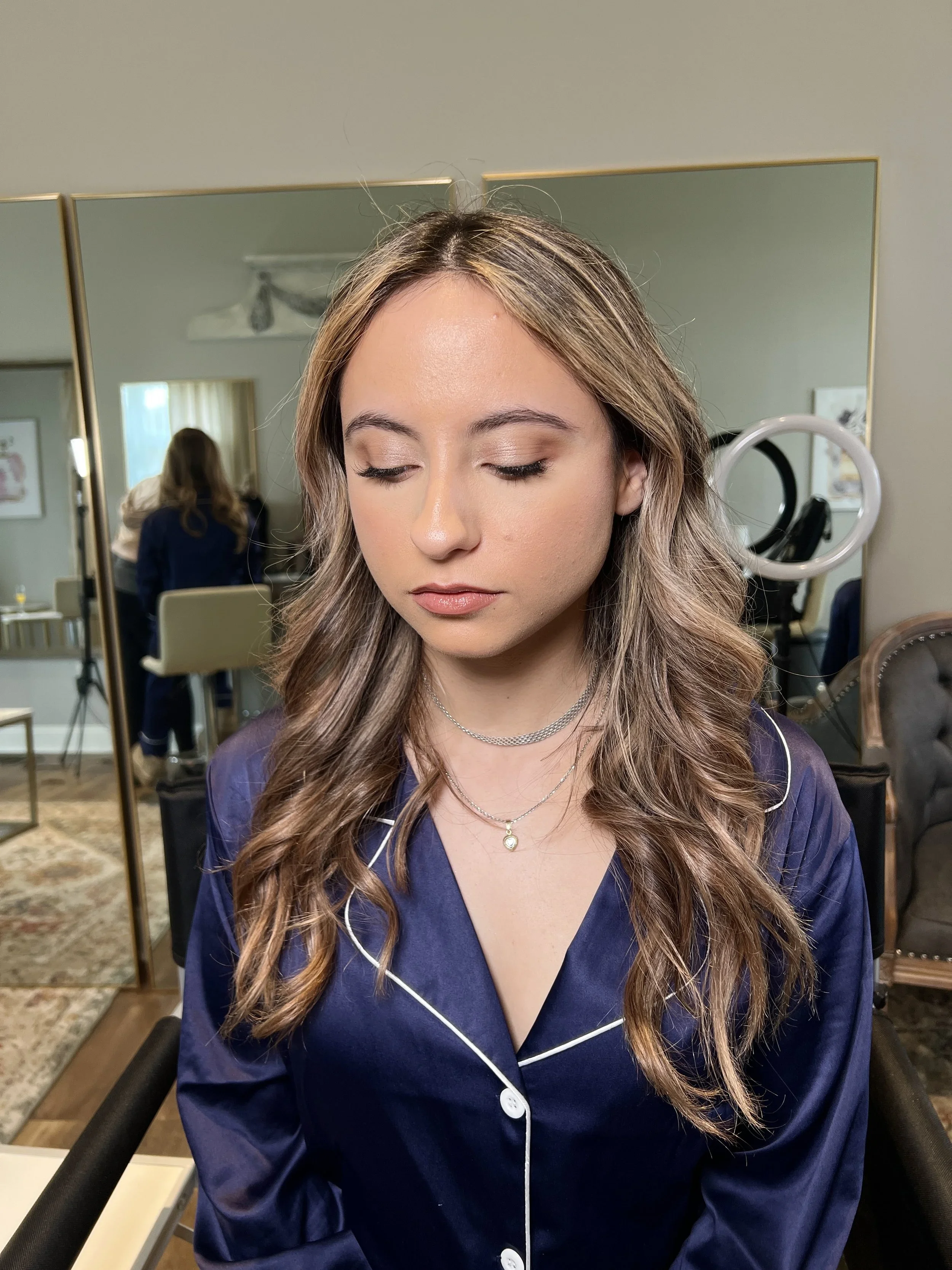 A woman with long wavy hair and light makeup wearing navy blue pajamas with white piping, seated in a salon or studio with mirrors and a ring light in the background.