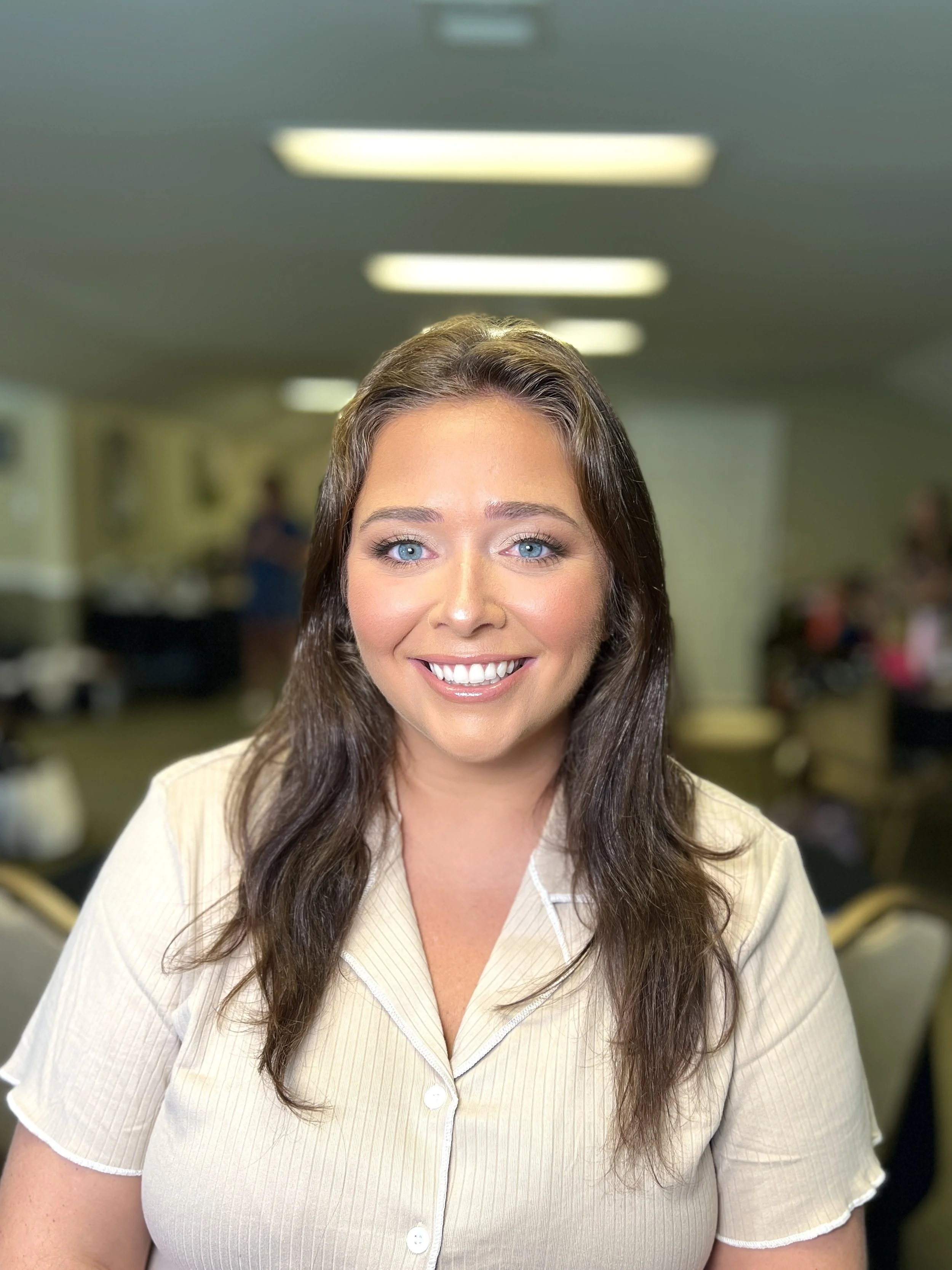 A smiling woman with long brown hair and blue eyes wearing a light-colored shirt in an indoor setting with blurred background and ceiling lights.
