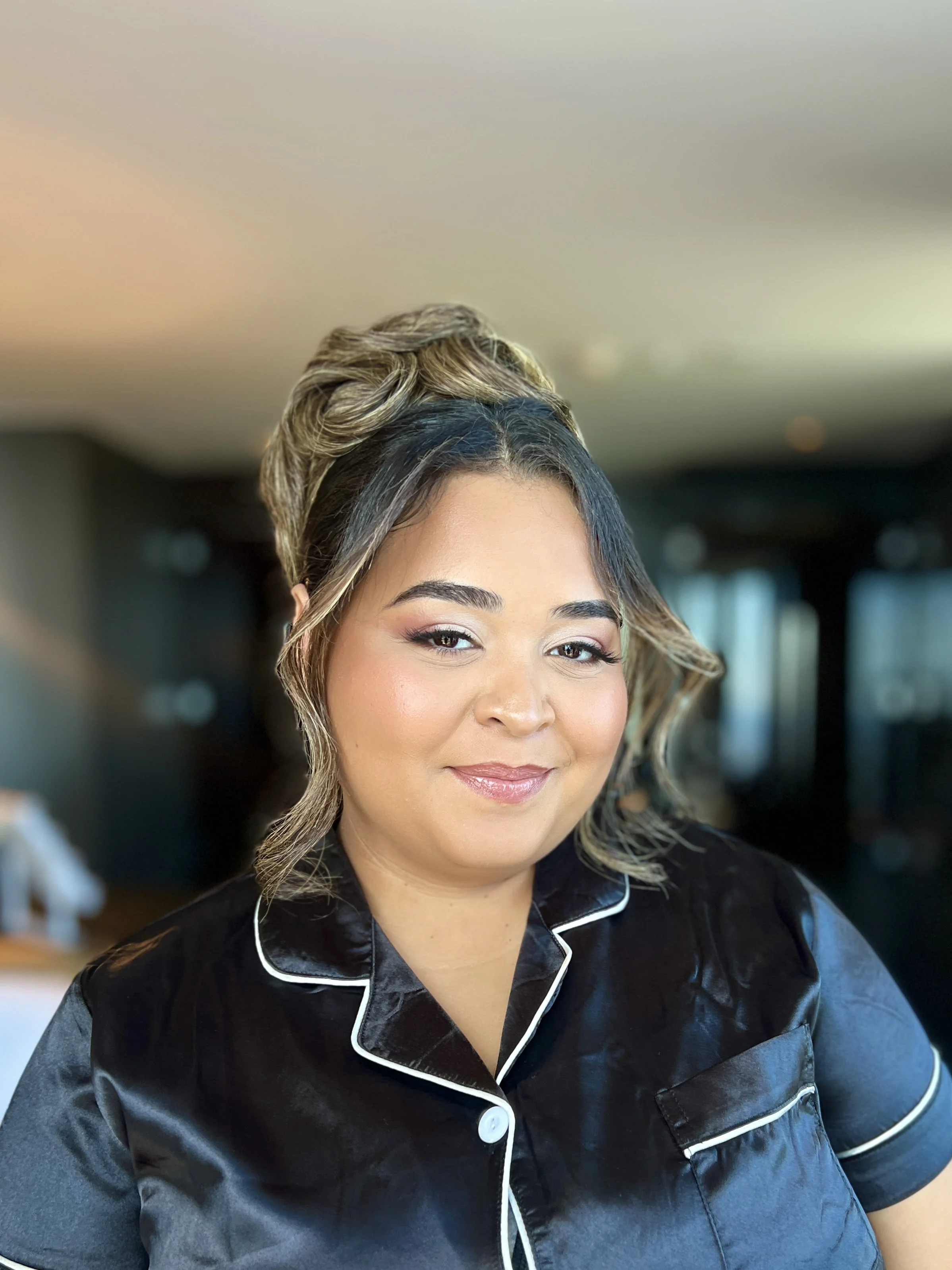 A woman with styled hair and light makeup smiling at the camera, wearing a black satin pajama top with white piping.