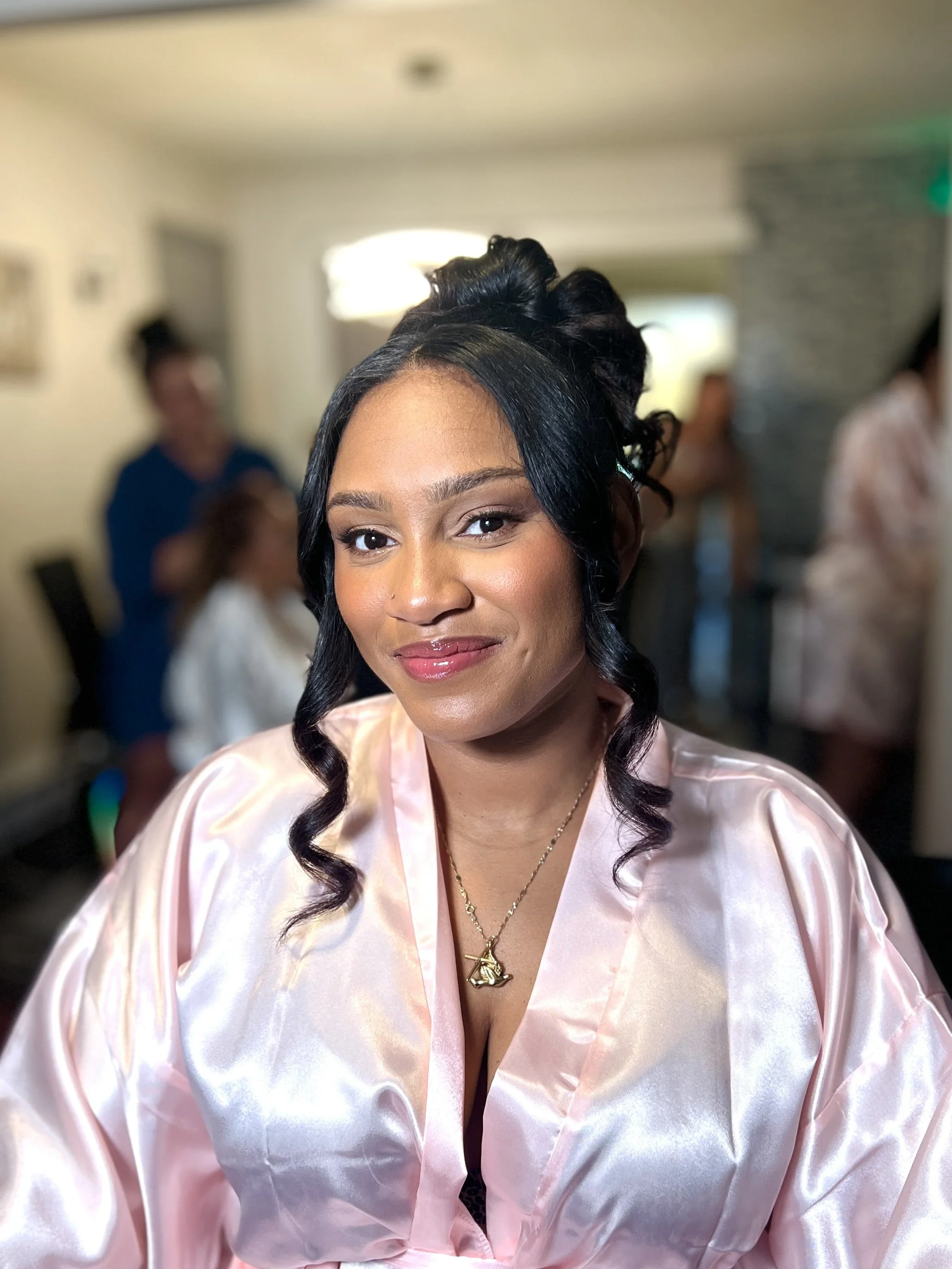 A smiling woman with dark hair styled in curls, wearing a light pink satin robe, and gold jewelry, inside a room with blurred background of people and a stone wall.