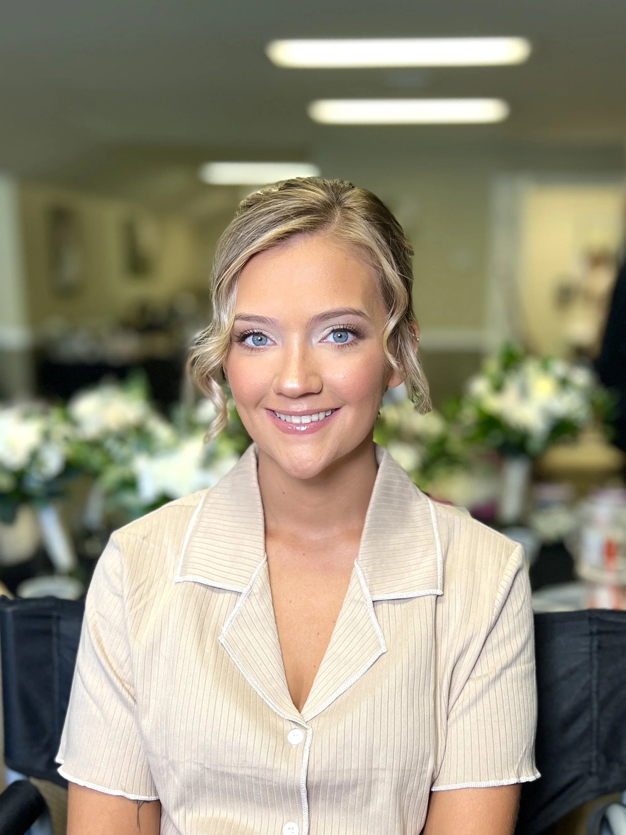 A smiling woman with blue eyes and blonde, slightly curled hair in a beige collared shirt, with floral arrangements in the background.