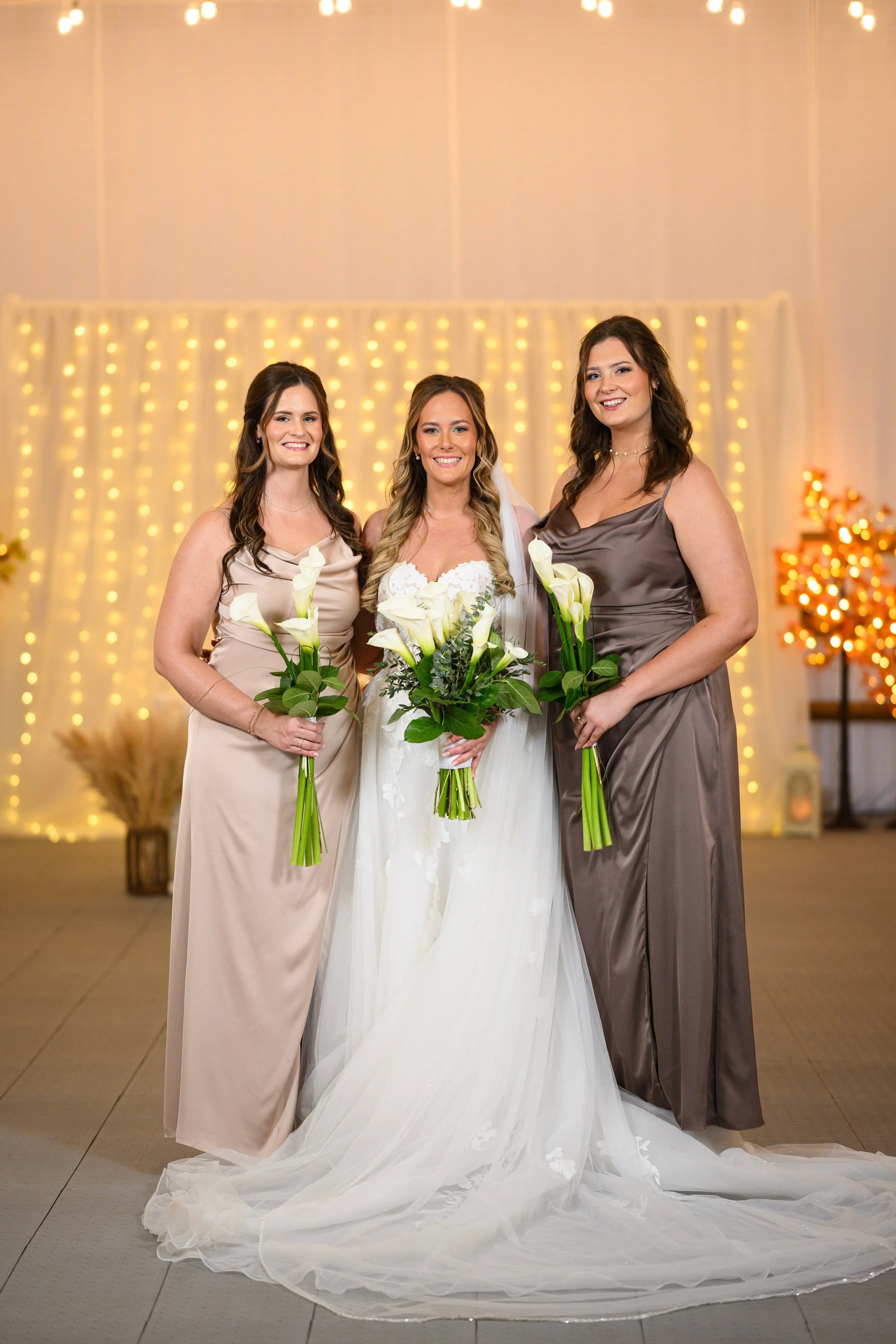 Three women, including a bride in a white wedding dress, standing together holding bouquets, in front of a curtain of warm string lights, with decorative trees and plants in the background.