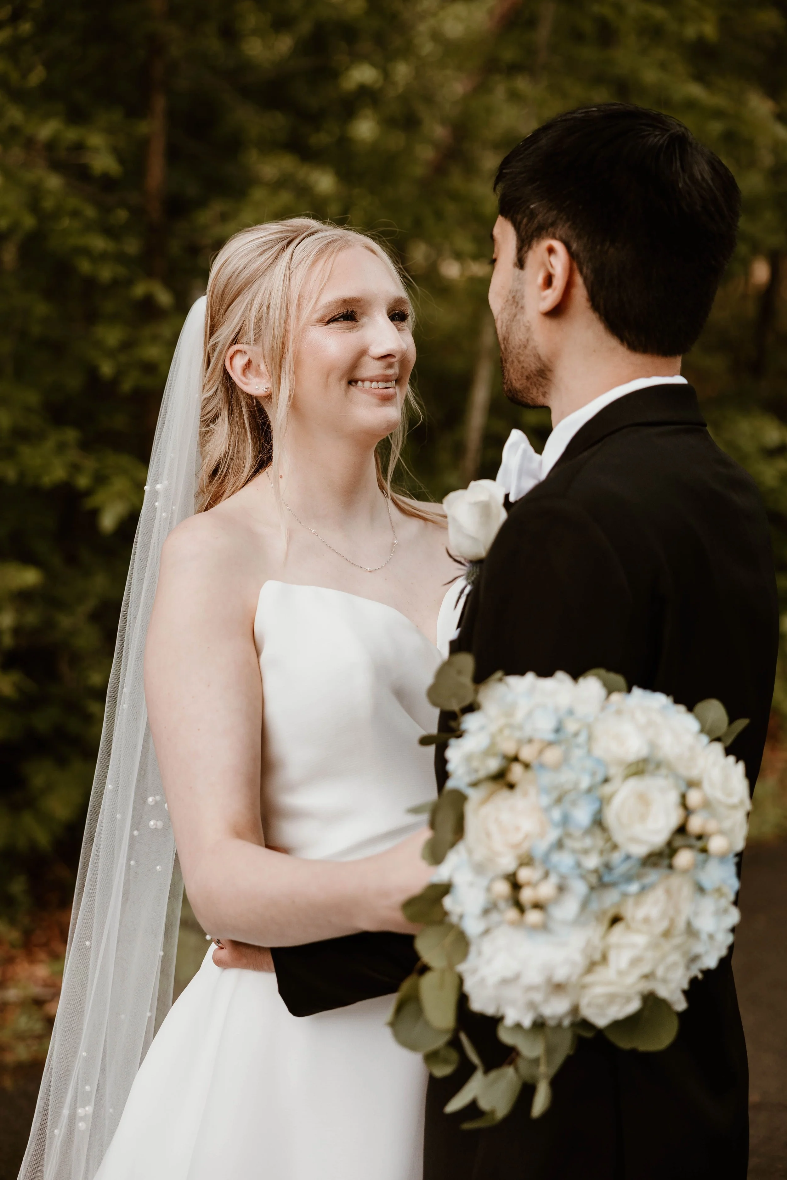 A bride and groom facing each other, smiling, outdoors, bride in white wedding dress with veil holding a bouquet, groom in black tuxedo with white boutonniere.