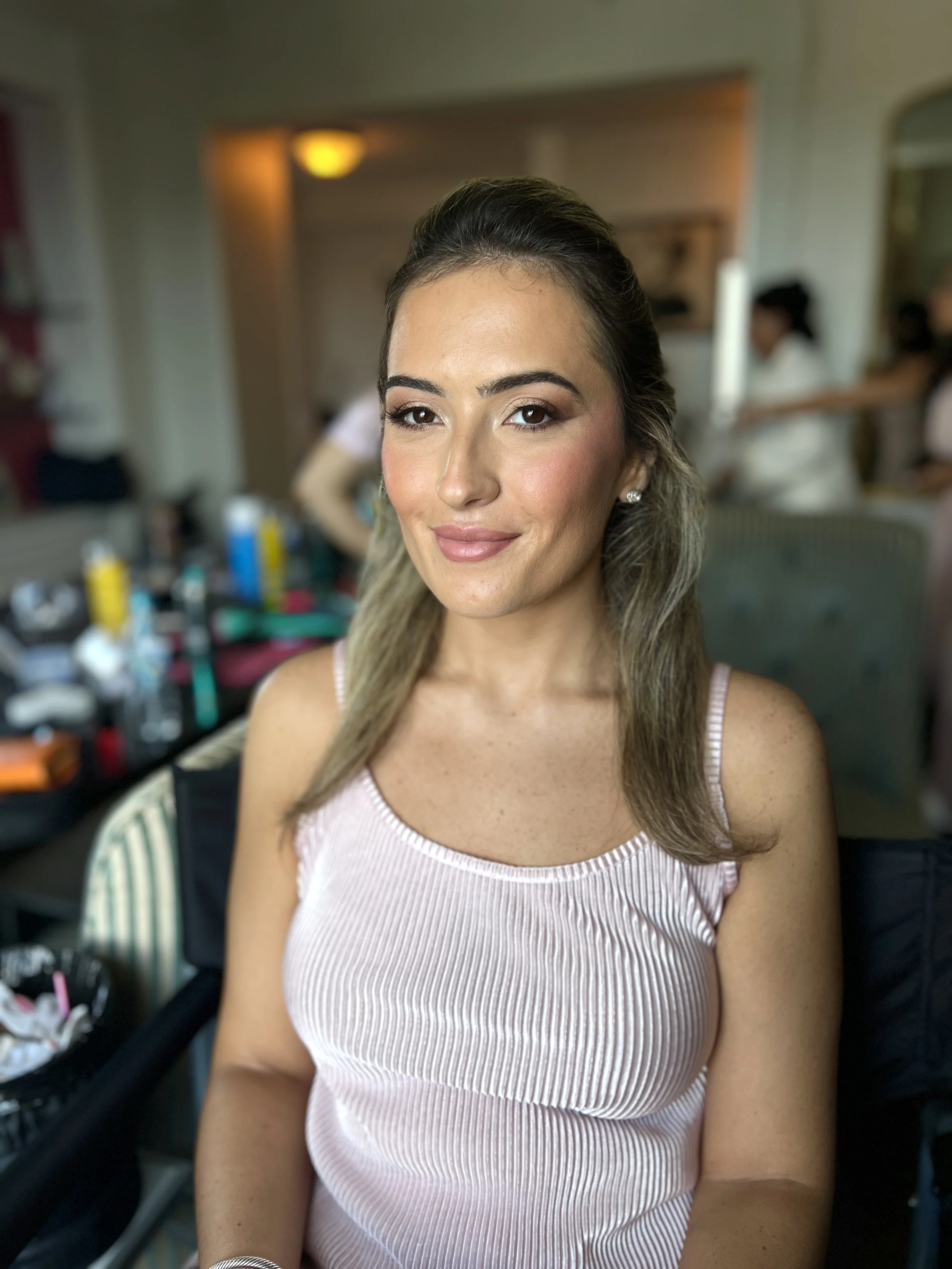 A woman with light skin and long, wavy brown hair, wearing a pink sleeveless top, sitting in a room with a blurred background of people and makeup supplies.