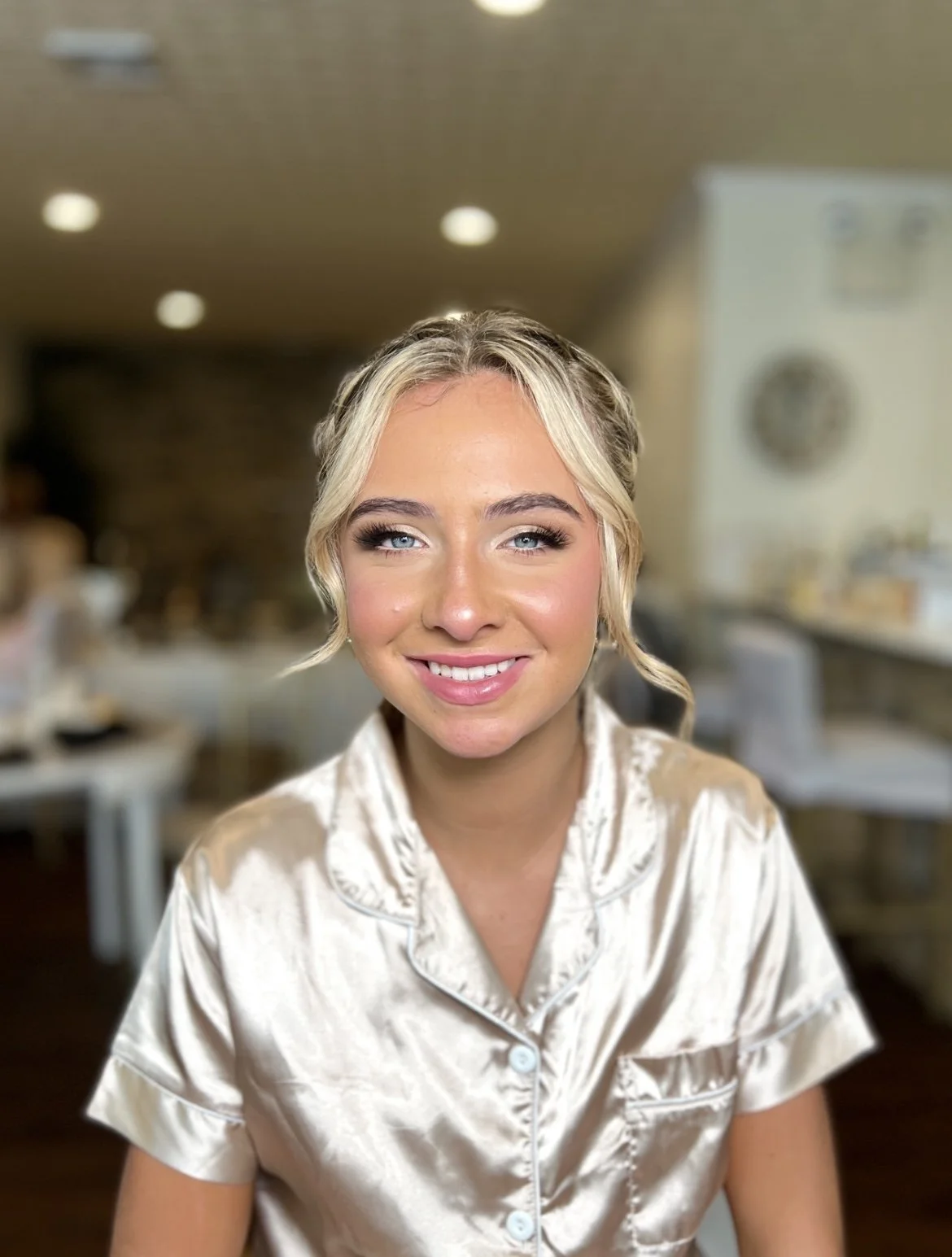 A young woman with blonde hair styled in loose waves, smiling, in a cream-colored silk pajama top, seated in a cozy indoor setting with warm lighting and blurred background.