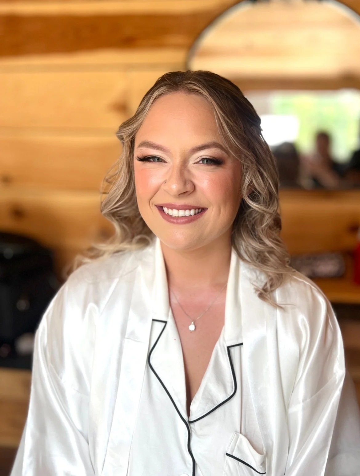 A woman with blonde curly hair, smiling, wearing a white satin pajama top with black piping, in a wooden room with a mirror in the background.