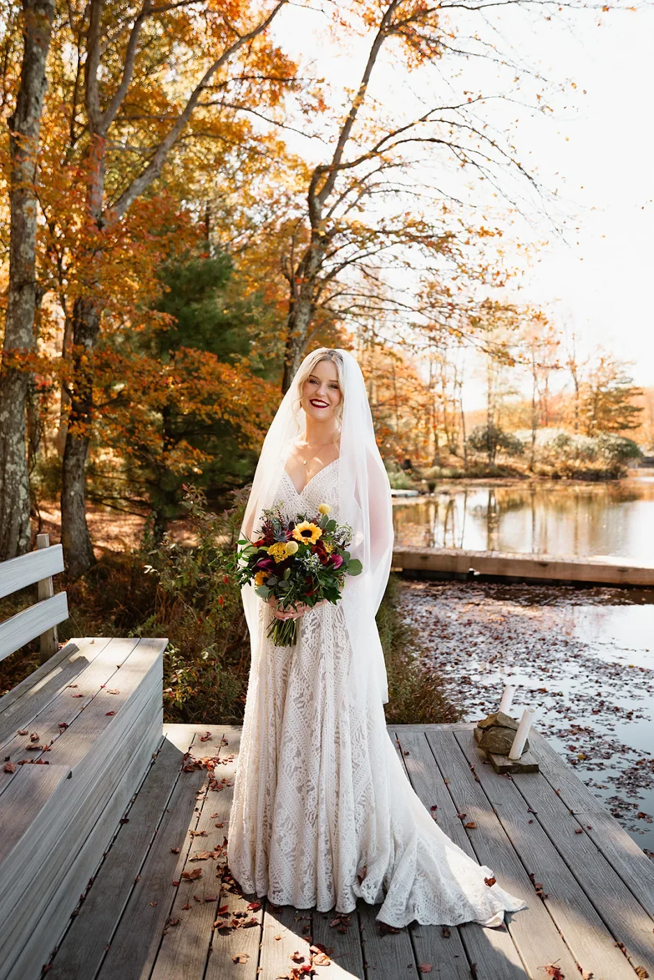 A bride in a white lace wedding dress holding a bouquet of sunflowers and colorful flowers, standing on a wooden dock by a lake, with trees displaying autumn foliage in the background.