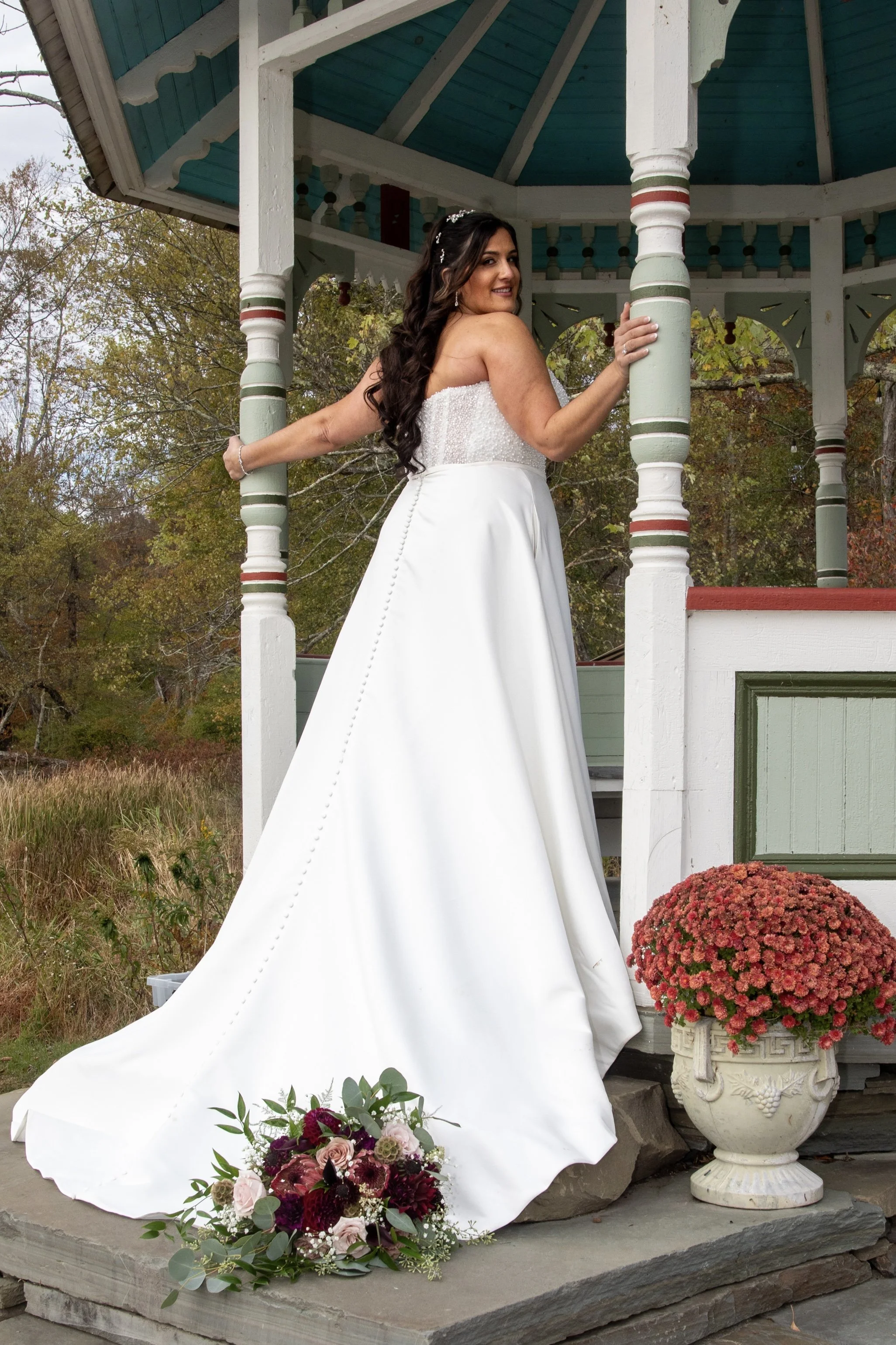 A bride in a white wedding dress standing on a porch, holding onto a column, with a bouquet of flowers on the ground nearby, and a large red potted plant beside her, during autumn.