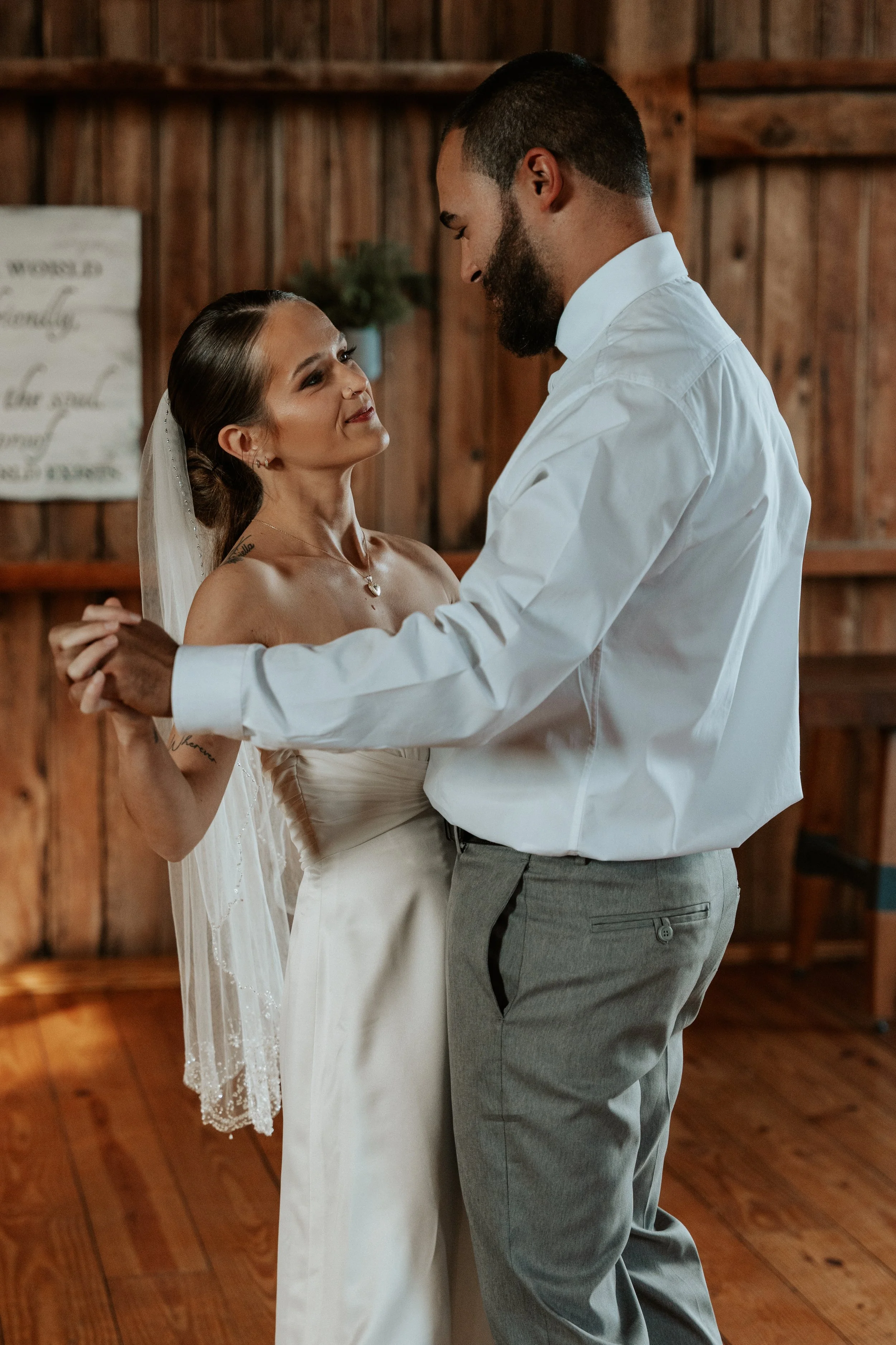 A bride and groom dance closely indoors with wooden walls in the background, the bride wearing a slip dress and veil, and the groom in a white shirt and gray pants.
