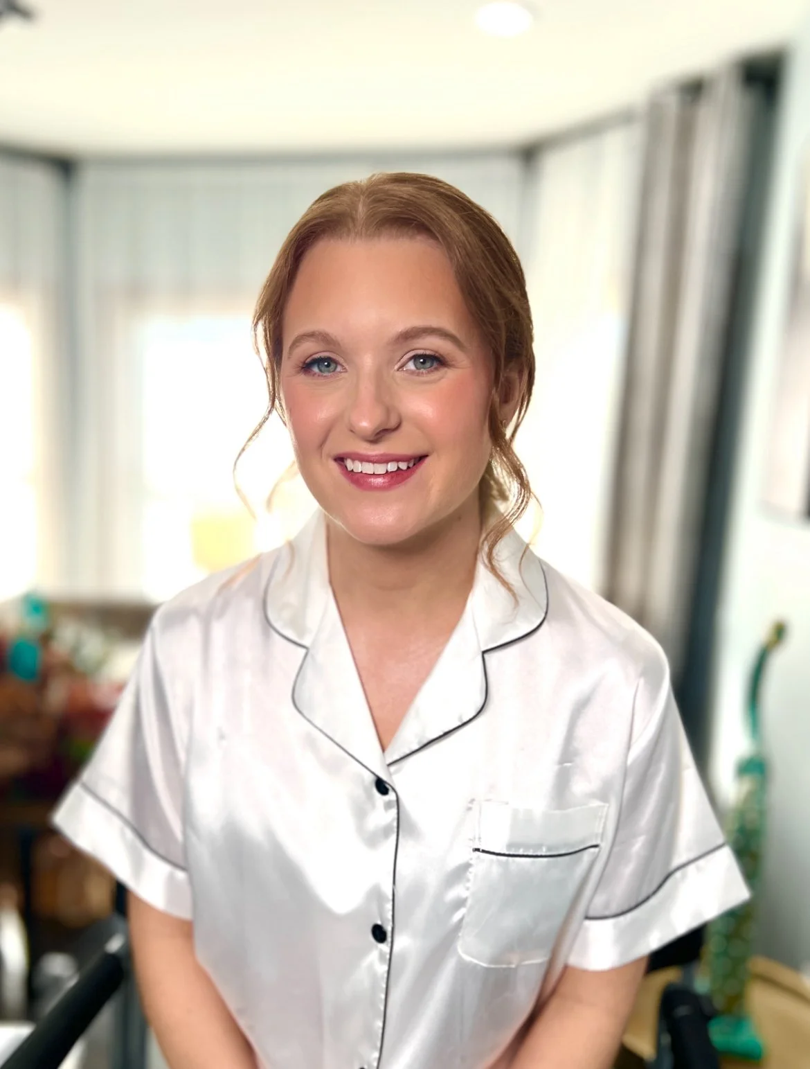 A woman with red hair in pajamas smiling at the camera in a bright room.
