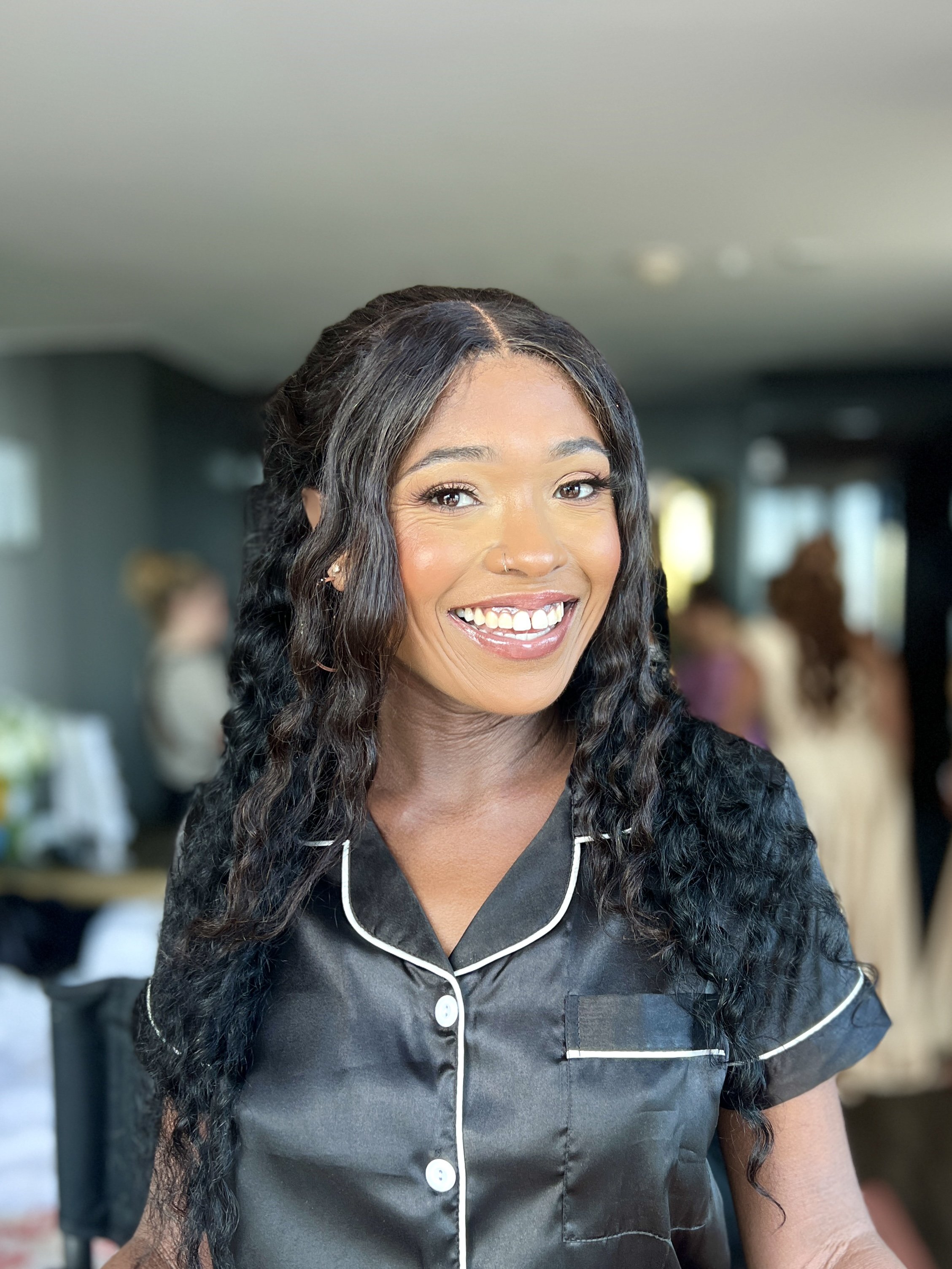 A smiling woman with long curly black hair, wearing a black satin pajama top with white piping, in a social setting with other blurred figures in the background.