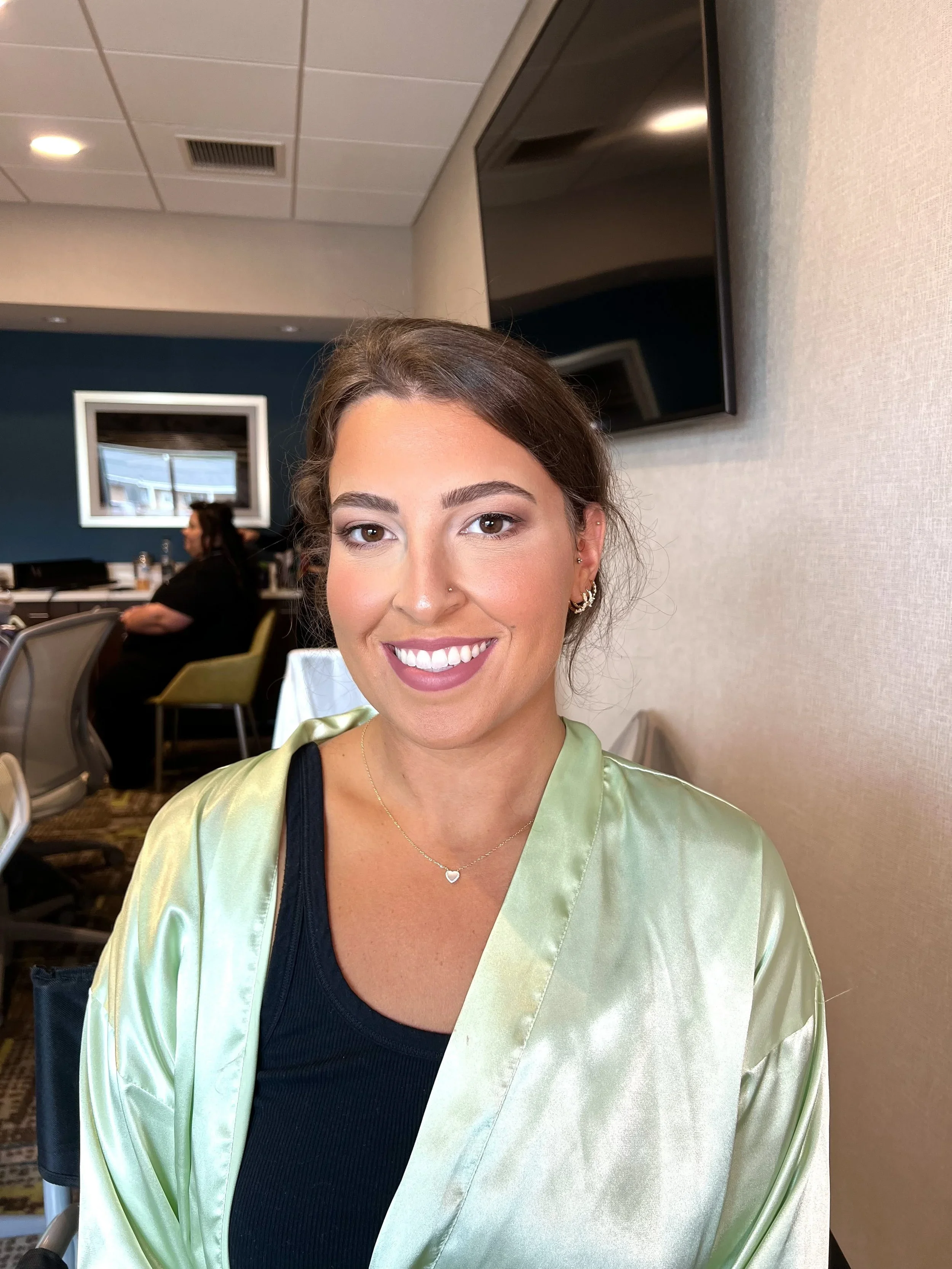 A smiling woman with brown hair and brown eyes, wearing a black top, a light green satin jacket, gold earrings, a necklace with a small heart pendant, and a nose piercing, sitting indoors in a room with beige walls and a large TV screen behind her.