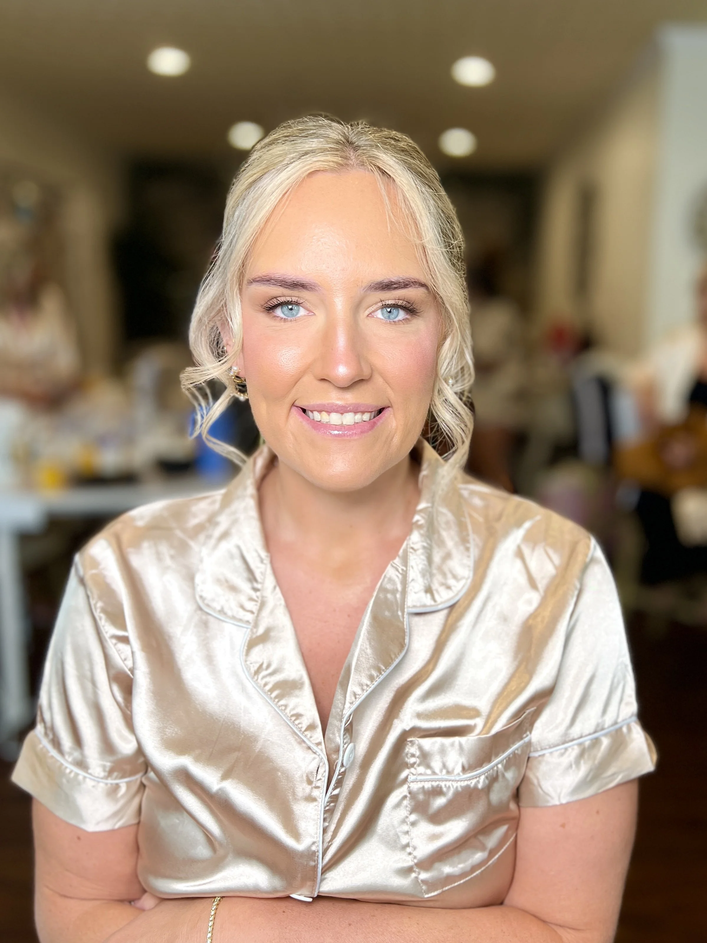 A woman with blonde hair and blue eyes smiling, wearing a satin beige button-up top, sitting in a warmly lit indoor setting with blurred background.