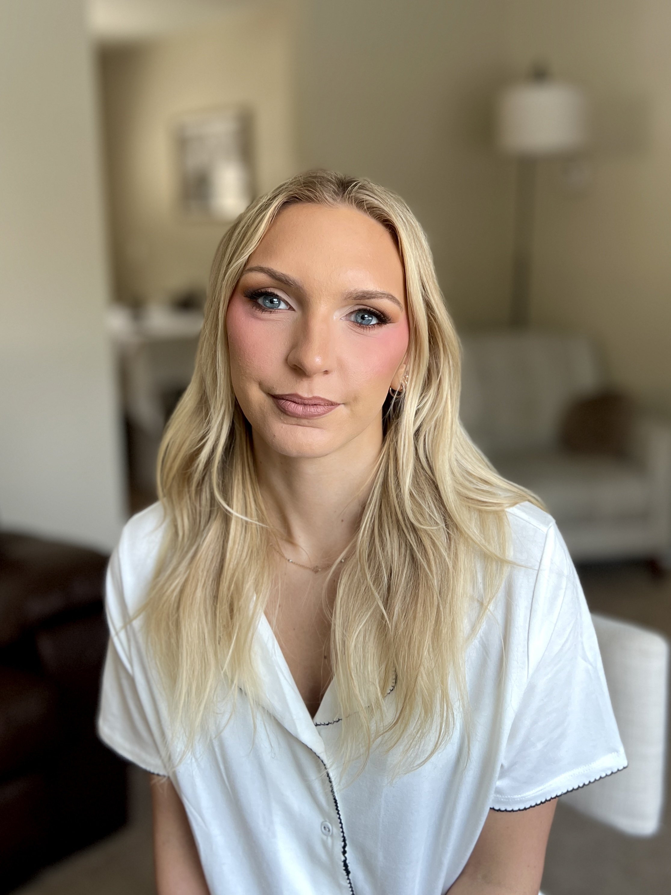 A young woman with long blonde hair and blue eyes, wearing a white collared shirt, sitting in a softly lit room with beige walls, a lamp, and a chair in the background.
