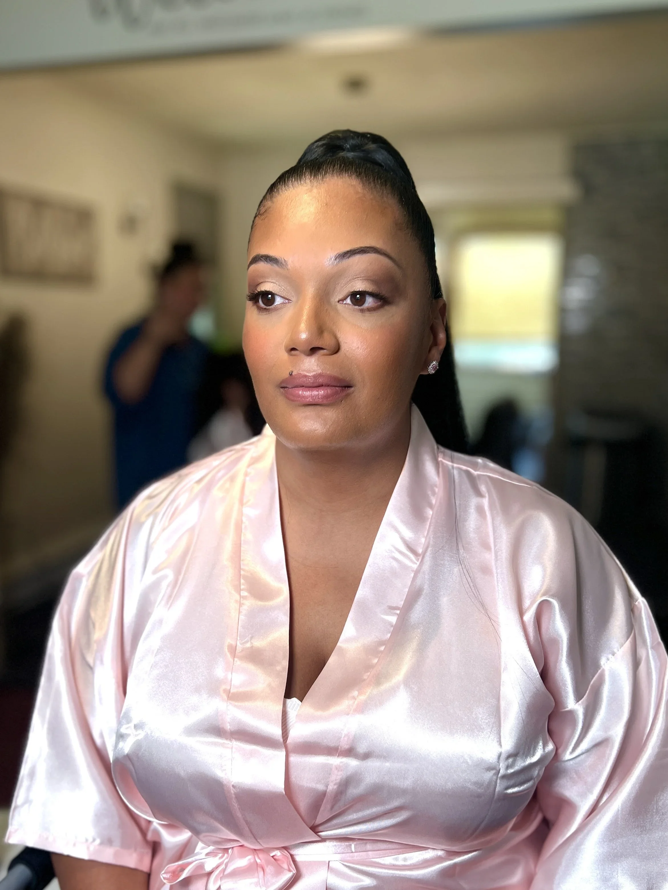 A woman with makeup and earrings wearing a pink satin robe, sitting indoors with a blurred background.
