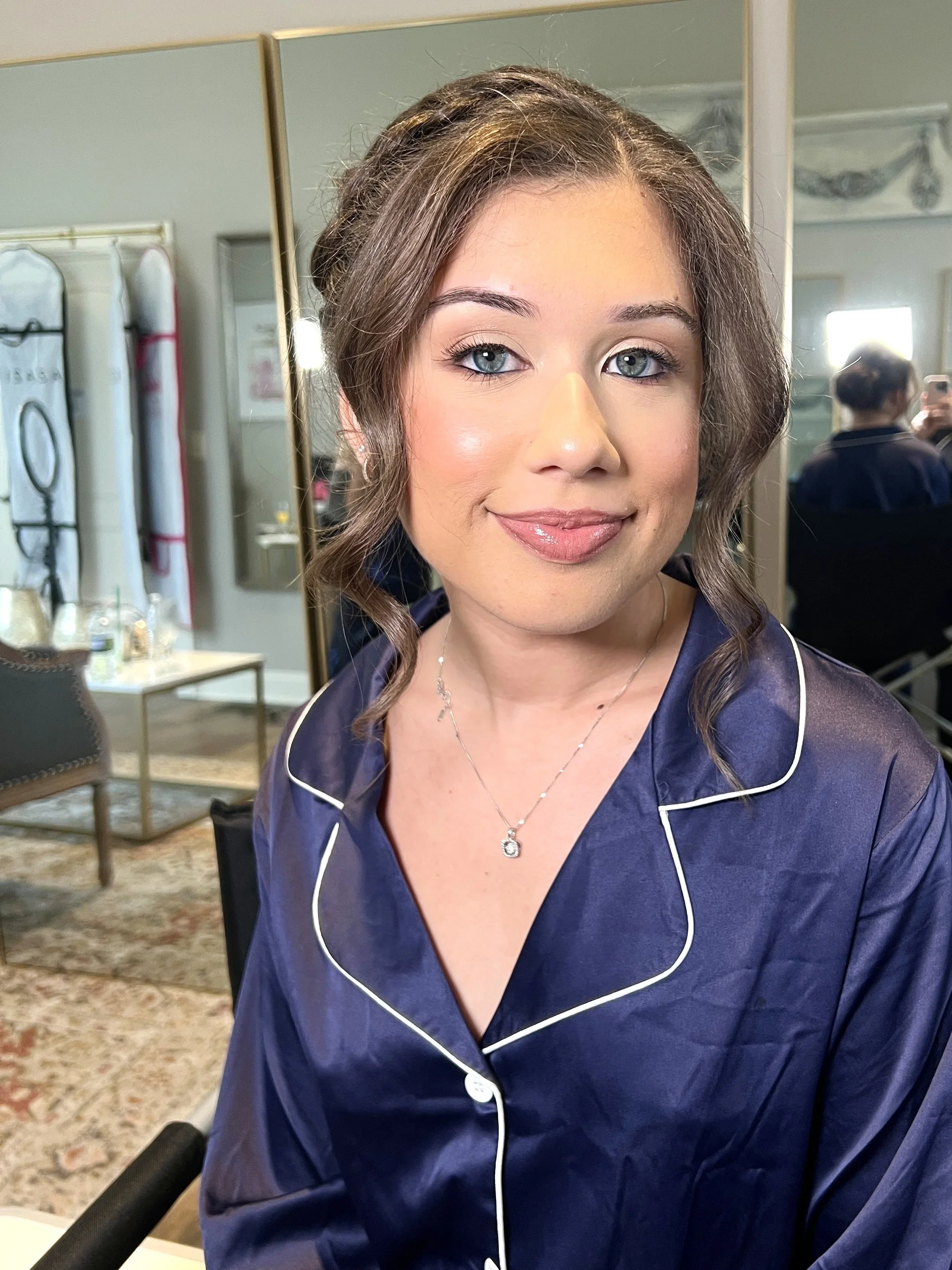 A woman with blue eyes, wearing a navy blue satin pajama top with white piping and a delicate necklace, smiling at the camera in a well-lit room with mirrors, chairs, and a table in the background.