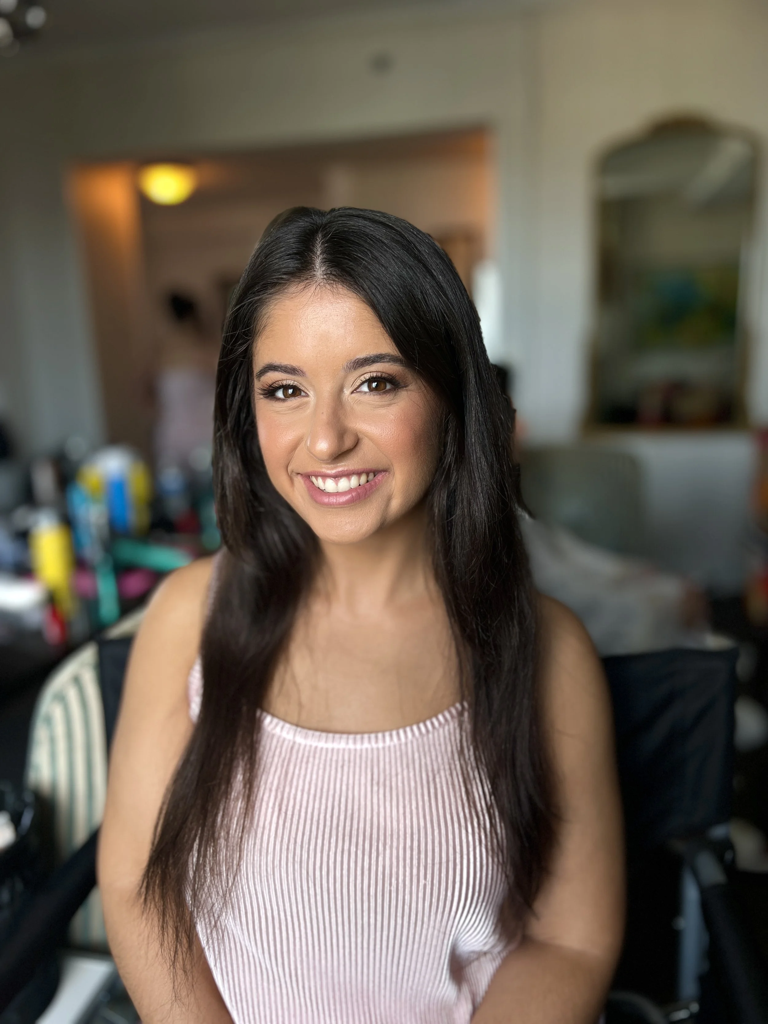 A woman with long dark hair and a pink sleeveless top smiling indoors.