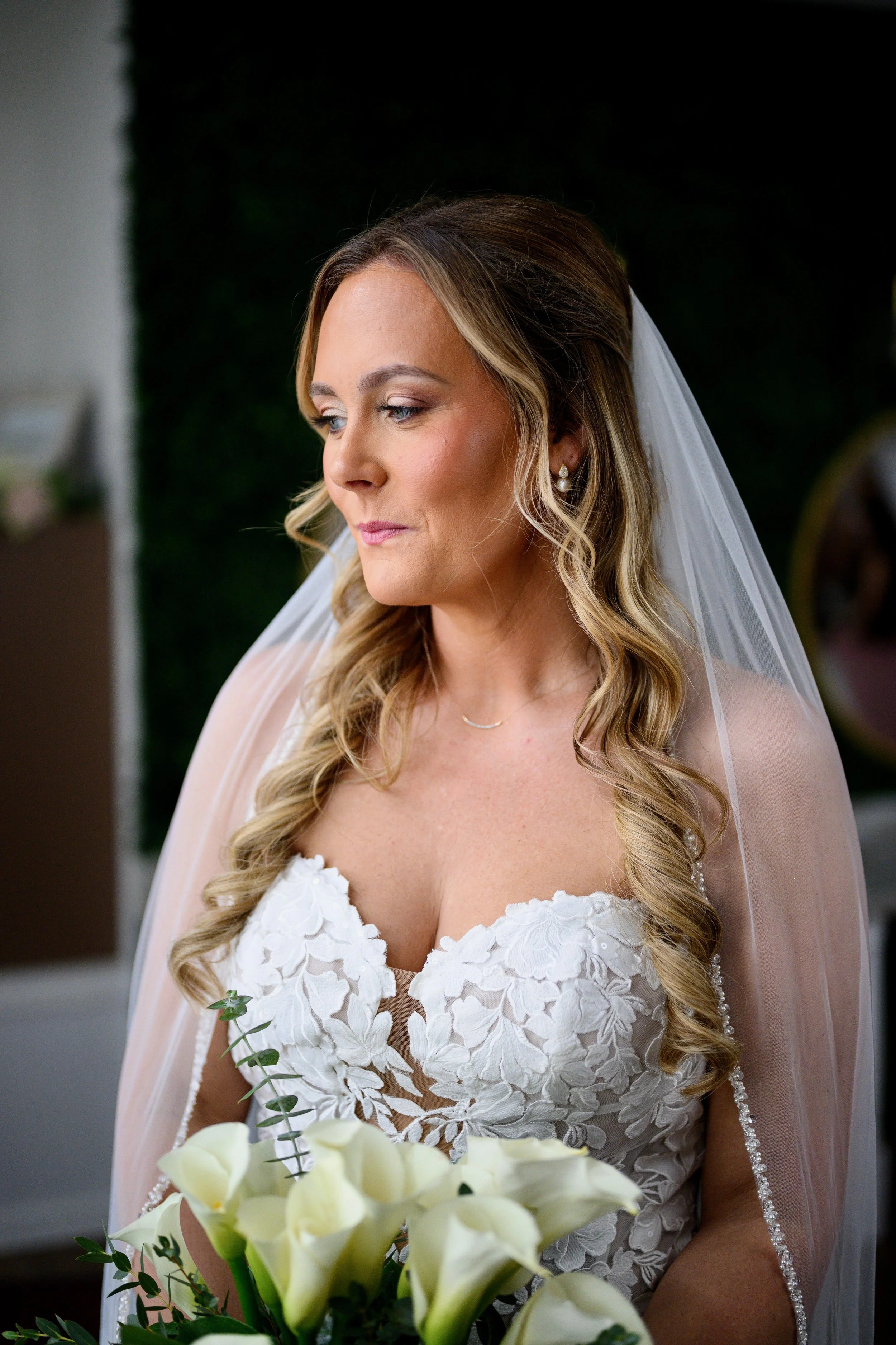 A bride with long wavy hair, wearing pearl earrings and a lace wedding dress with floral embroidery, holding a bouquet of white calla lilies, standing indoors with a dark background.