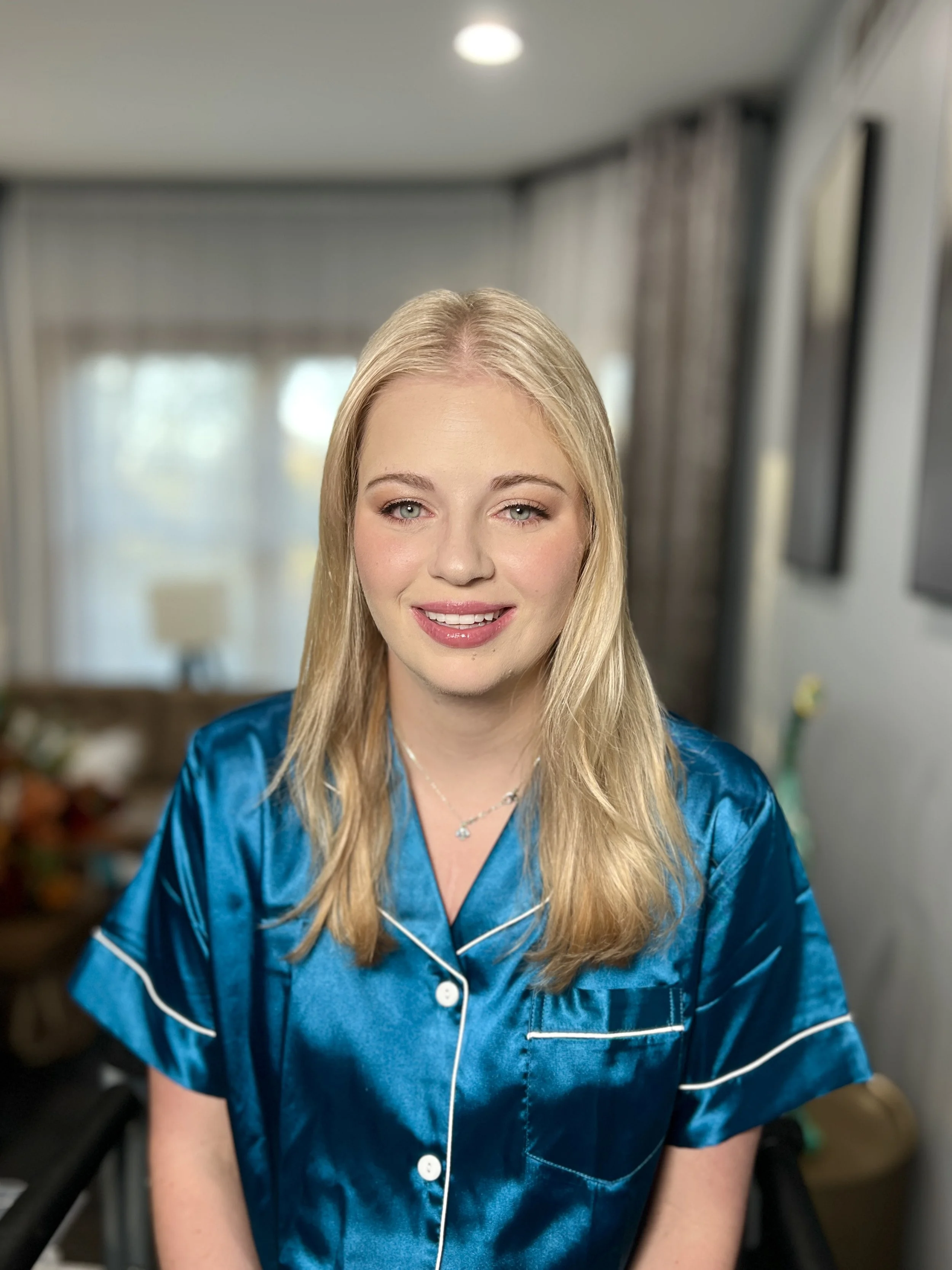 Blonde woman with makeup wearing a blue satin pajama top, sitting indoors with curtains and a window in the background.