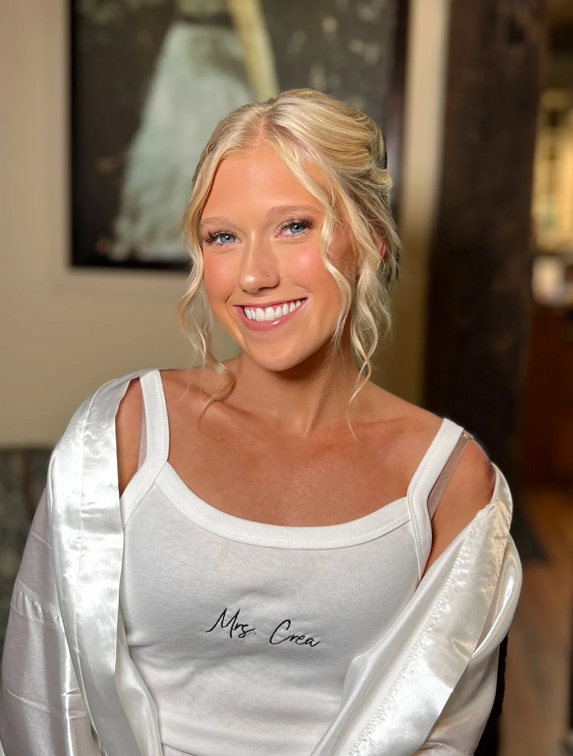 A smiling woman with blonde curly hair, wearing a white tank top with "Mrs..Orea" written on it, and a satin robe over her shoulders.