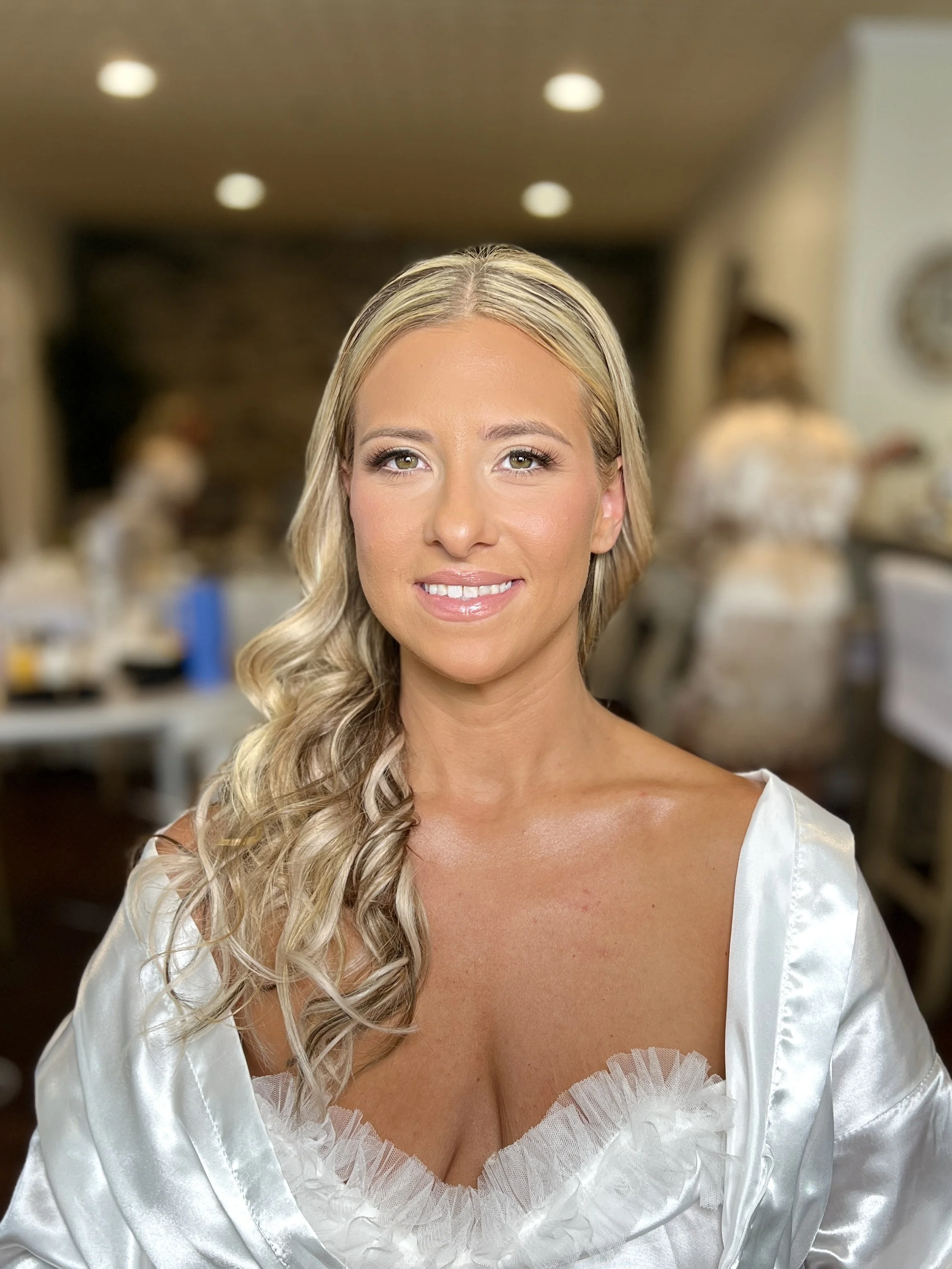 A blonde woman with styled hair and makeup, wearing a satin robe with lace trim, smiling at the camera inside a room with blurred background.