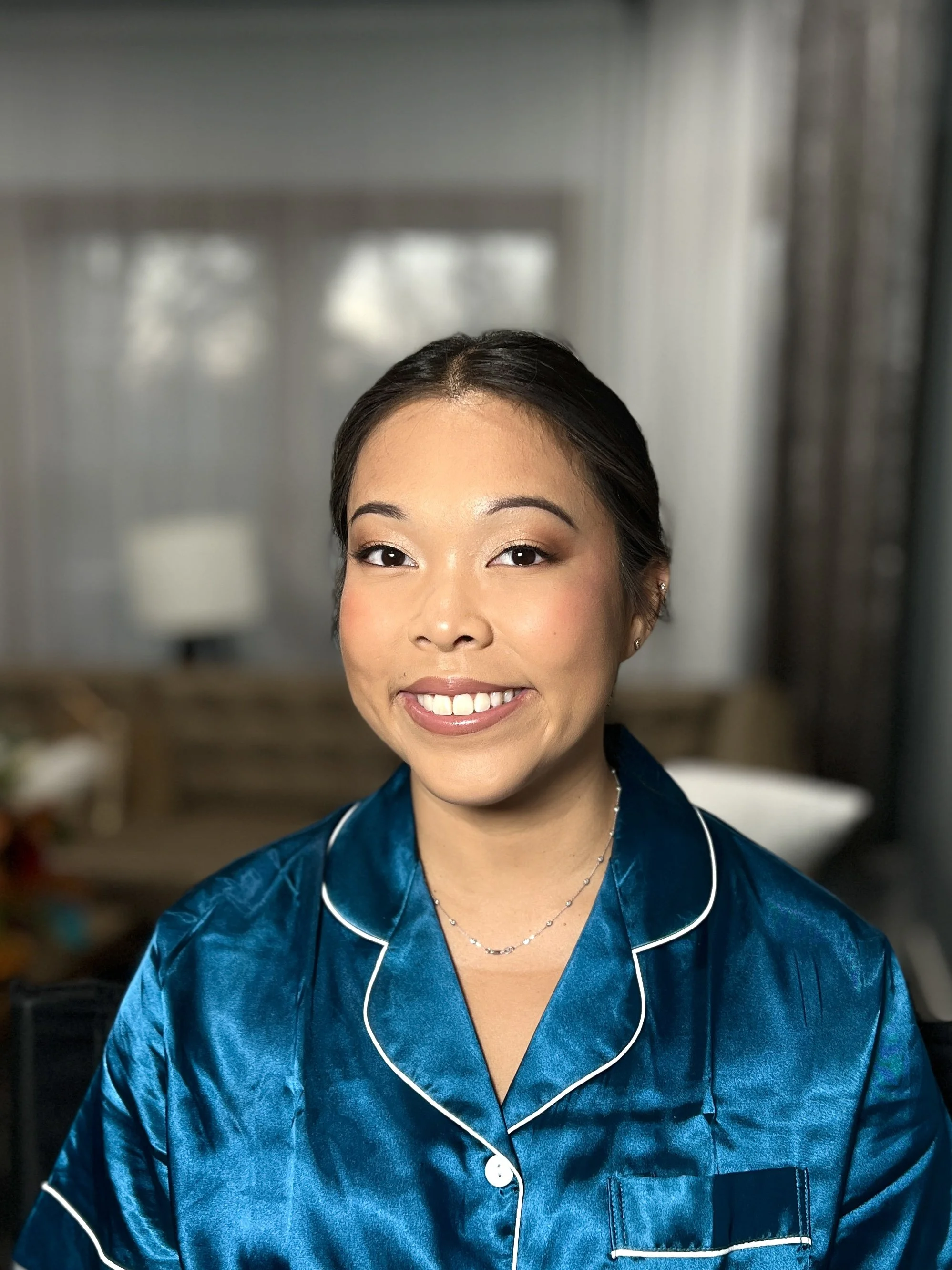 A woman smiling in a blue silk pajama top inside a room with light-colored curtains and a blurred background.