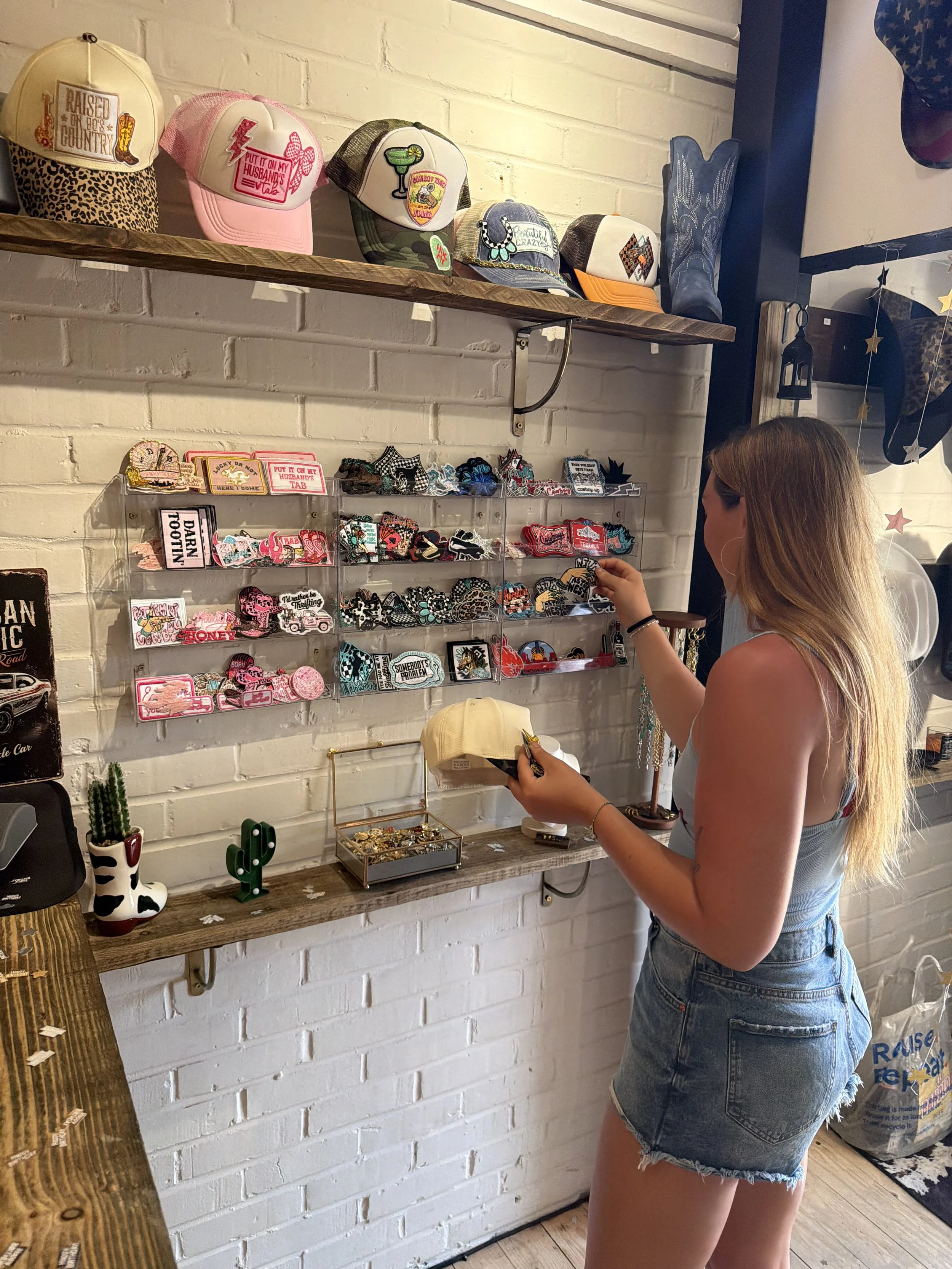 A woman with long hair, wearing a denim cut-off shorts and a tank top, shopping for patches at a boutique. She is holding a patch in her hand, standing in front of a white brick wall with display shelves of patches and hats, and decorative items like cactus-shaped planters.