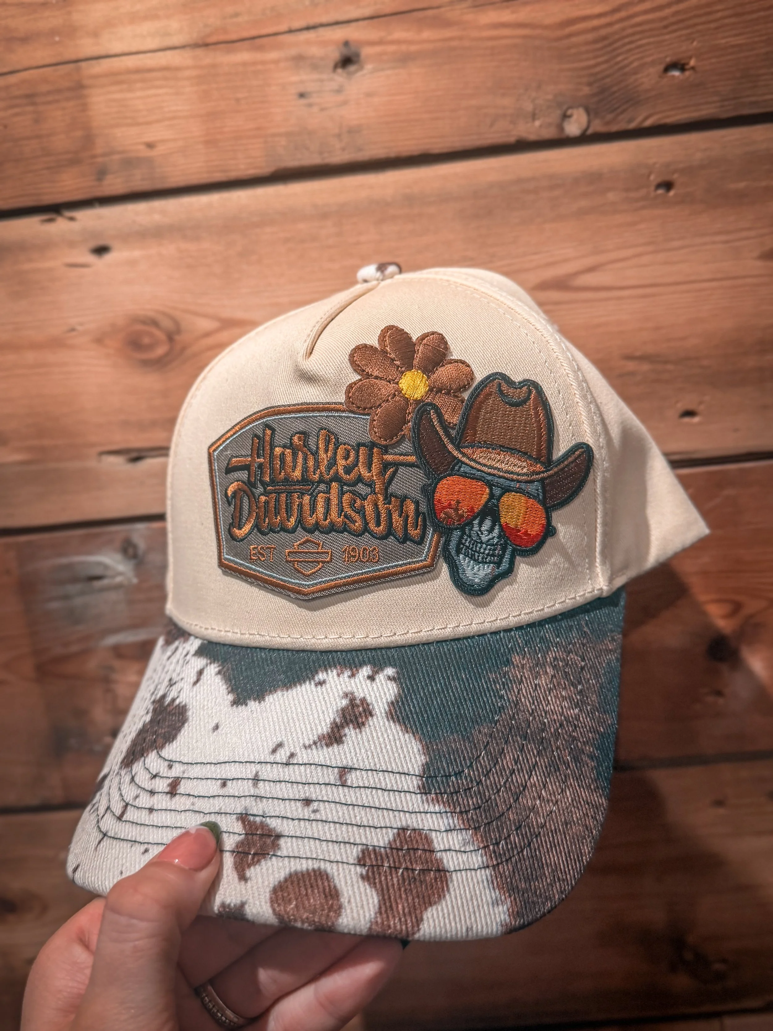 A person's hand holding a cowboy-themed Harley-Davidson cap with patches, on a wooden floor.