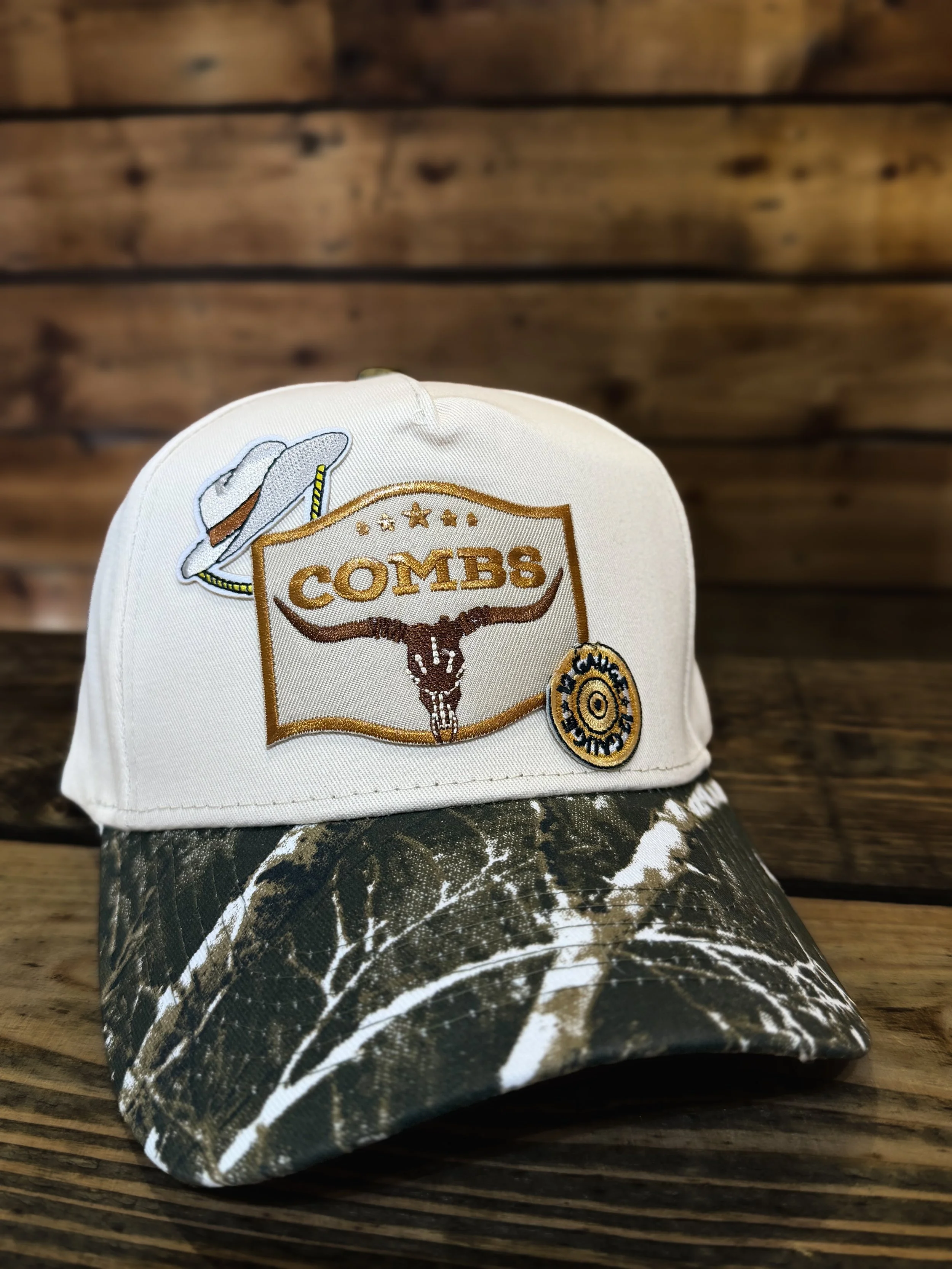 A white cap with embroidered patches of a hat, a longhorn skull, and a circular badge with a black and gold design, placed on a camouflage-patterned fabric, with a wooden wall in the background.