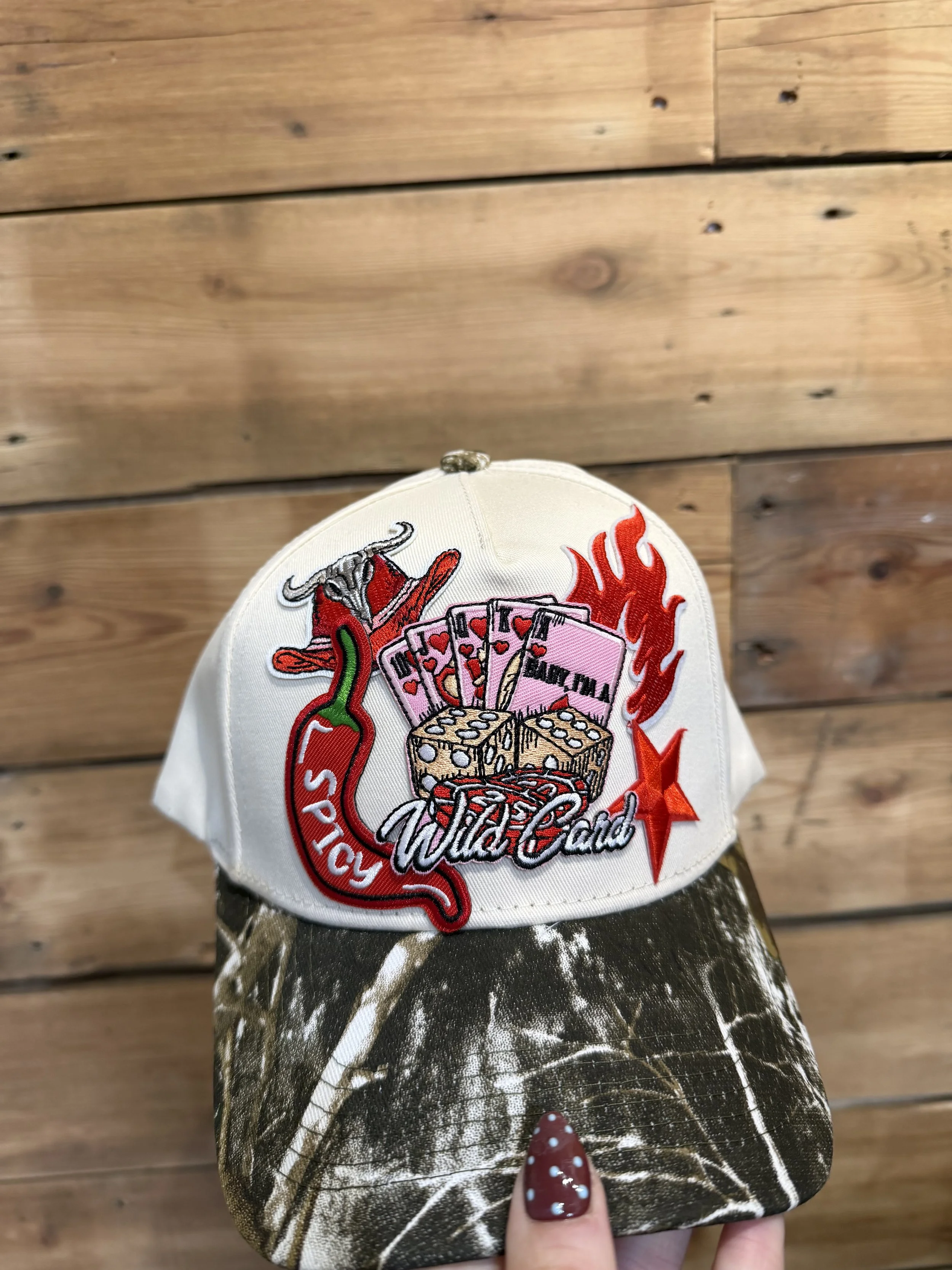 A white baseball cap with a colorful embroidered patch showing poker chips, a flaming chili pepper, and the word 'Wild Card', held by a person's hand with red and white polka dot painted fingernails, against a wooden floor background.