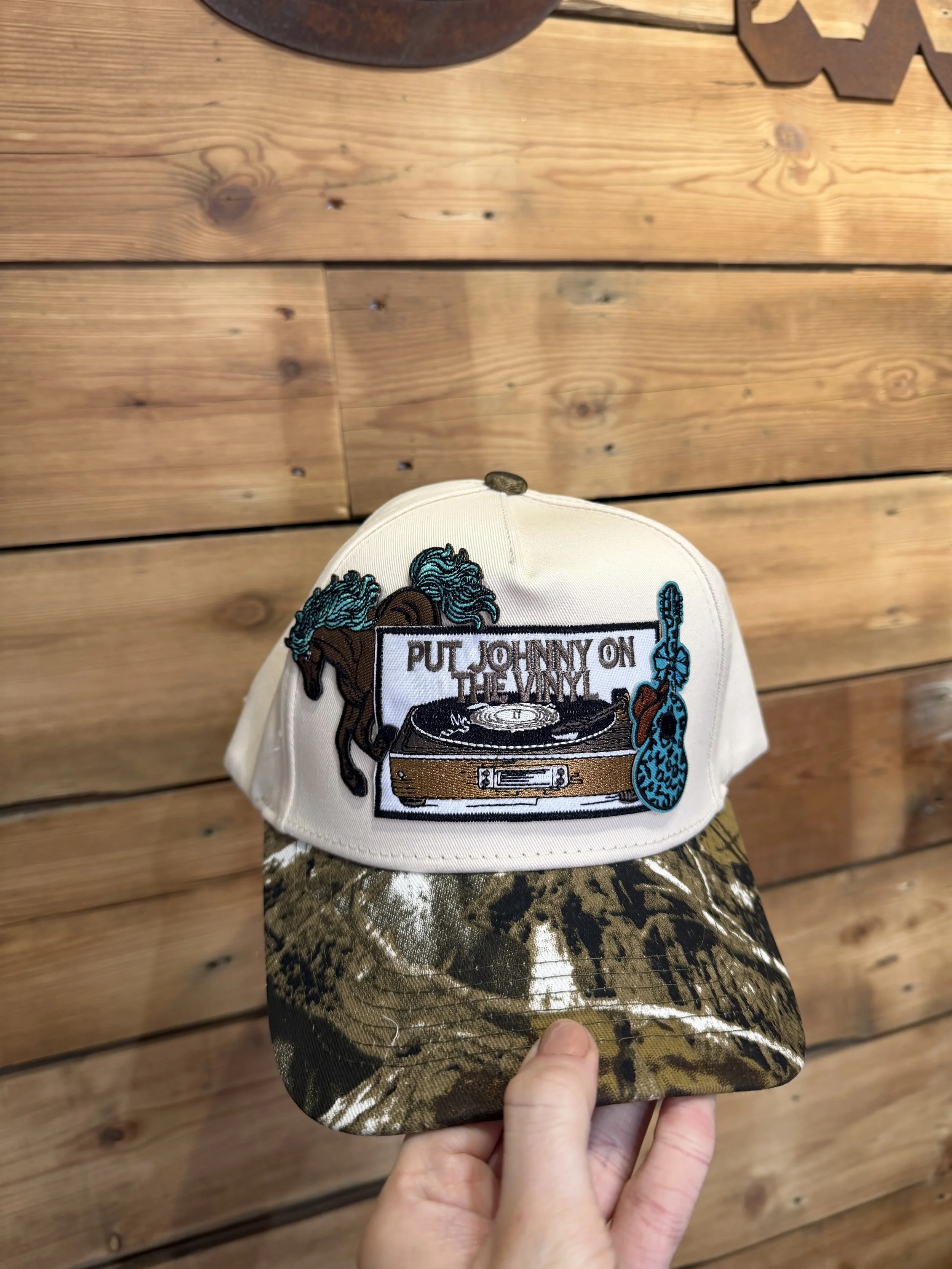 A baseball cap with a patch that says 'PUT JOHNNY ON THE VINYL' and features a vinyl record player, headphones, a horse, and a leopard.