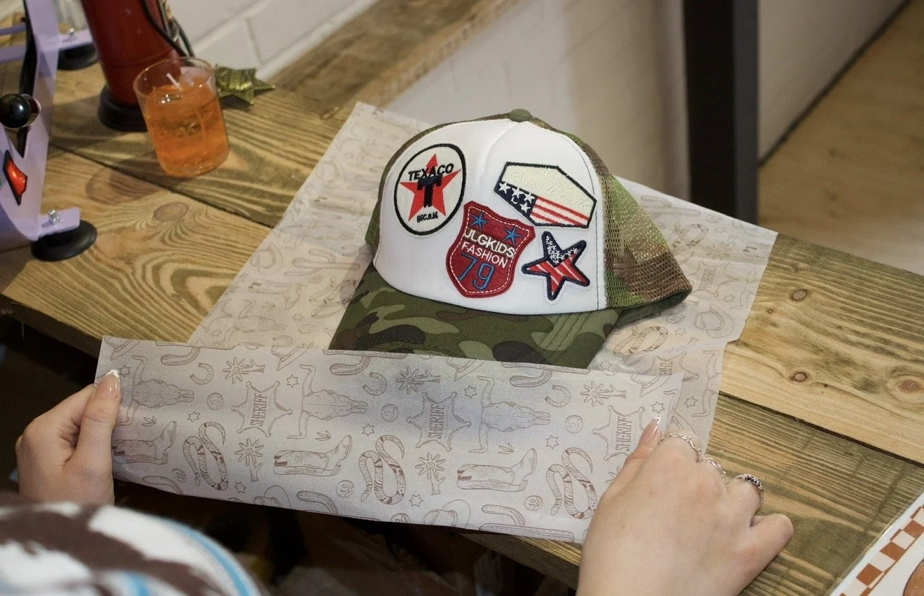 A camouflage baseball cap with patriotic patches on a wooden table. A hand is holding a sheet of paper with festive patterns beneath the cap.